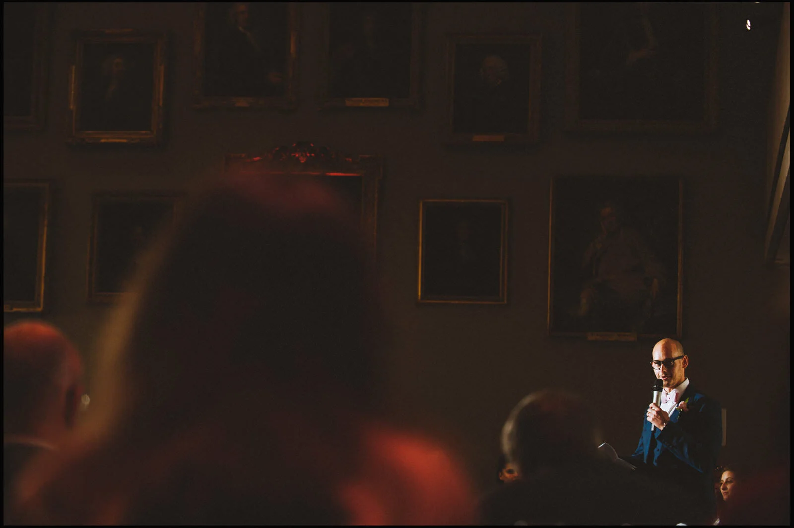 A man in a suit and glasses speaking into a microphone in a dimly lit room with framed portraits on the wall.