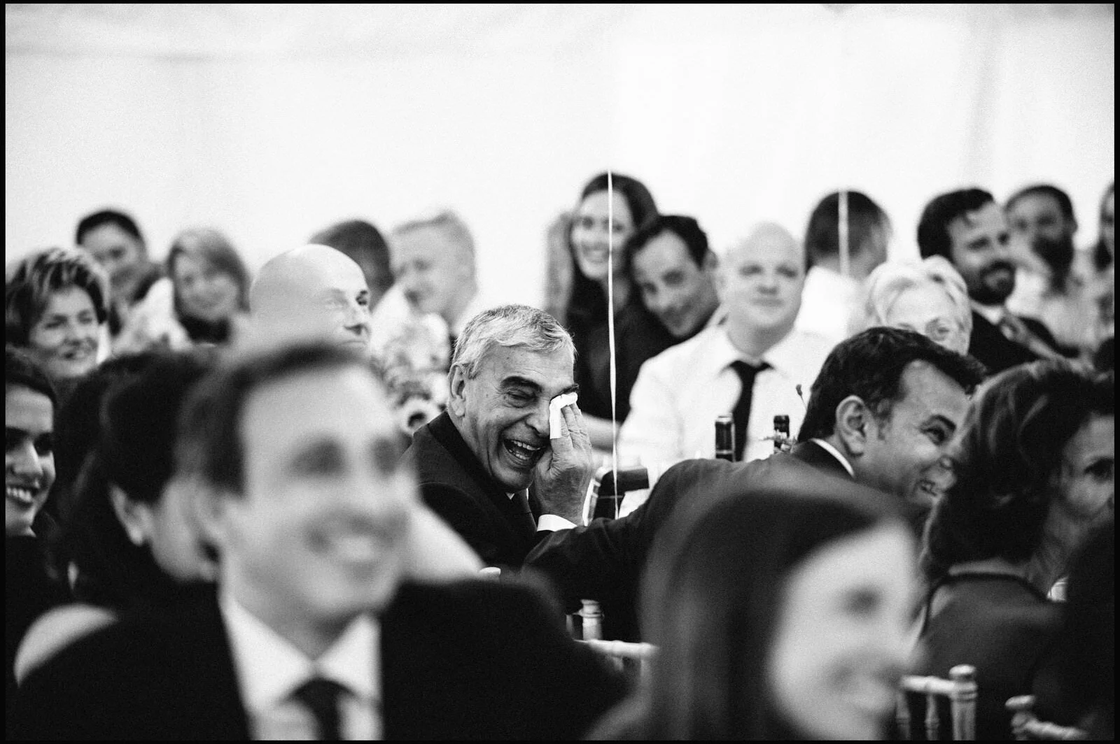 Black and white photo of people laughing and smiling at a gathering, with a man in the center wiping a tear or laughter from his face.