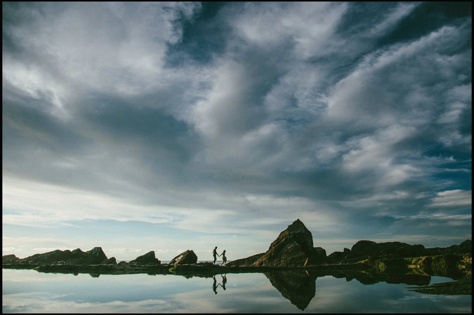 A couple holding hands walking on a rocky shoreline with a large rock formation, reflected in calm water, under a cloudy sky.