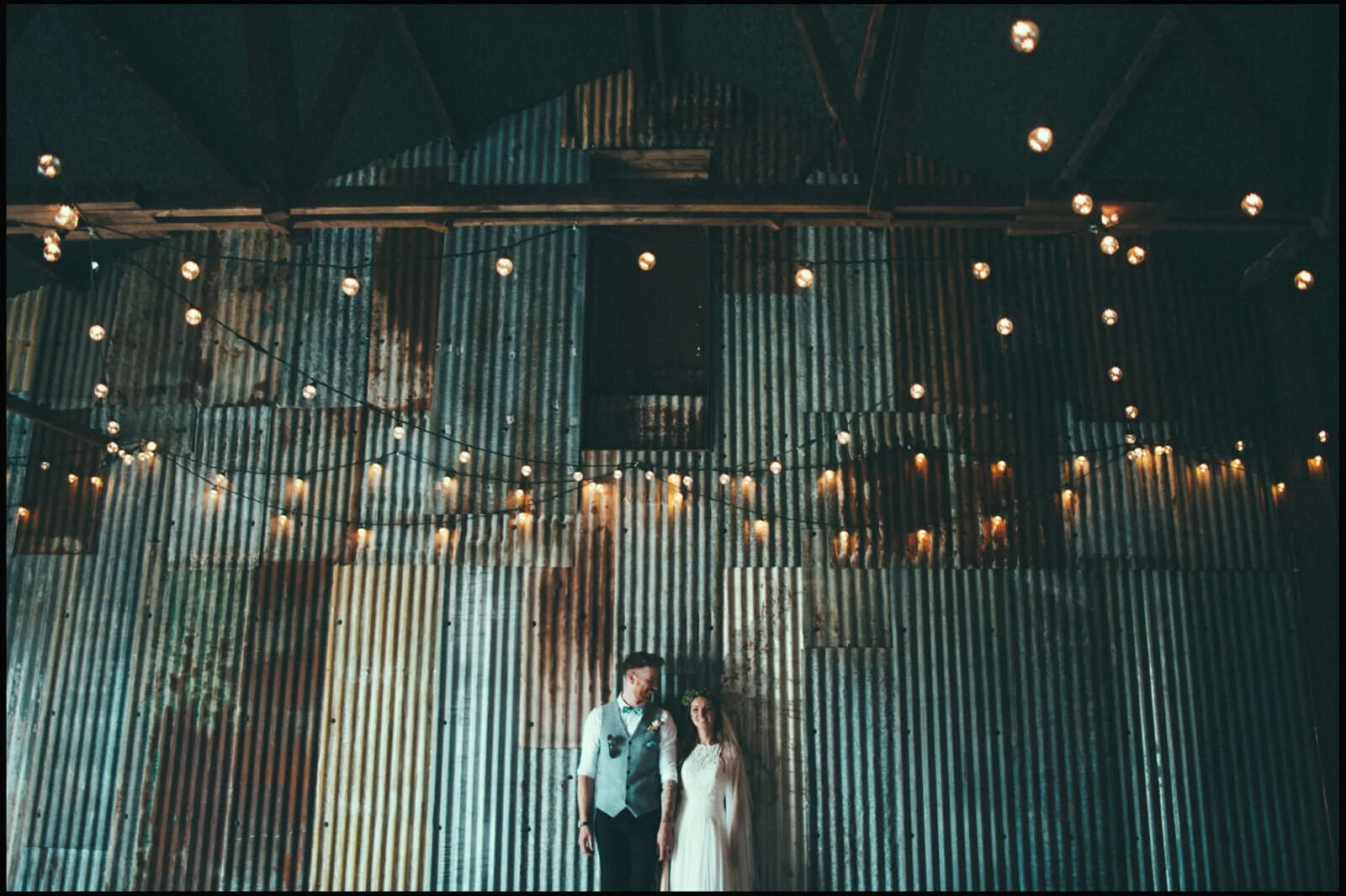 A bride and groom standing close together inside a rustic venue with corrugated metal walls and hanging string lights.