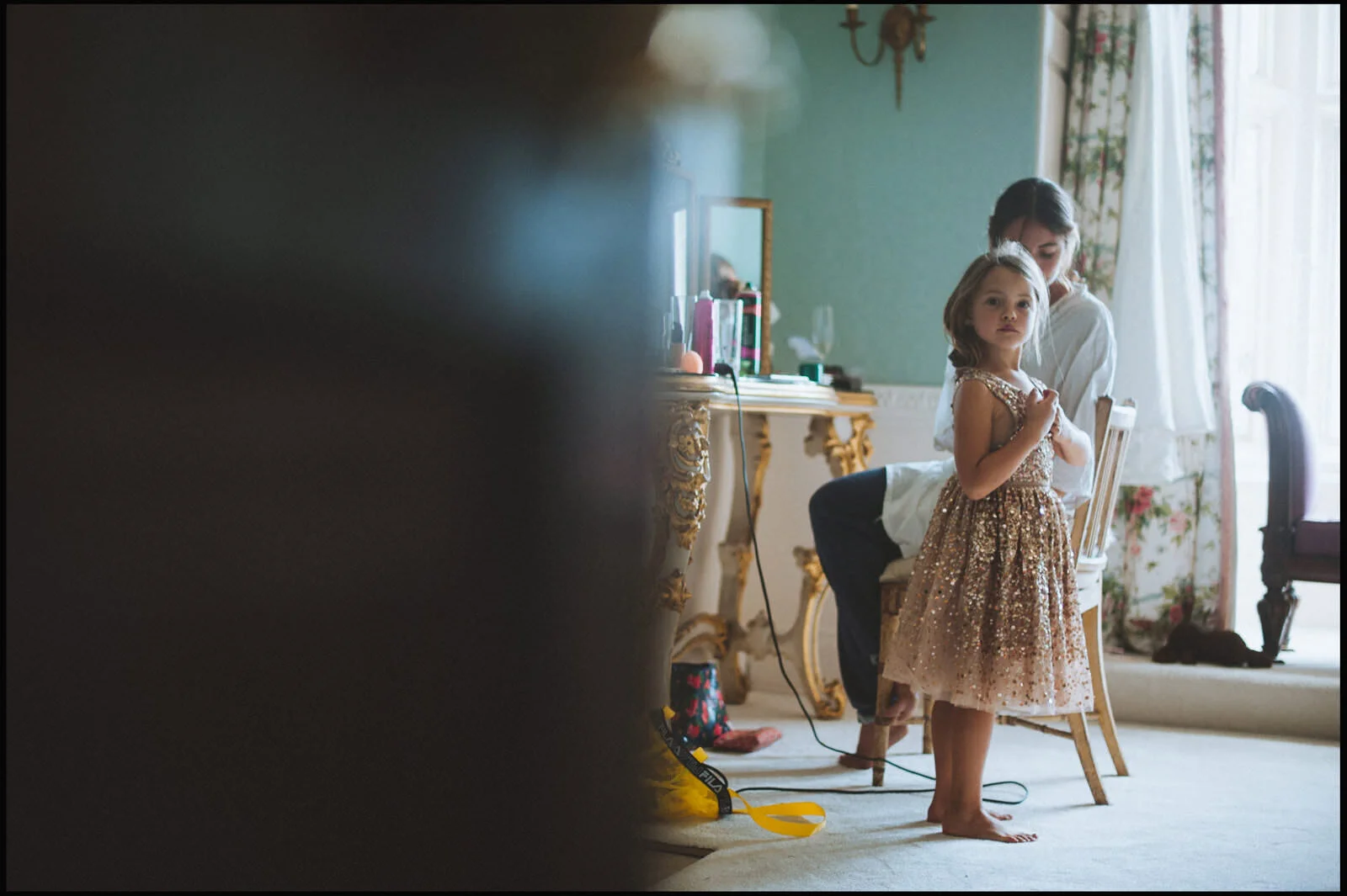 A young girl in a glittery gold dress standing in a room, with an adult woman behind her. The room has a vintage-style vanity with gold details, a mirror, and floral curtains. There is sunlight coming through a window.