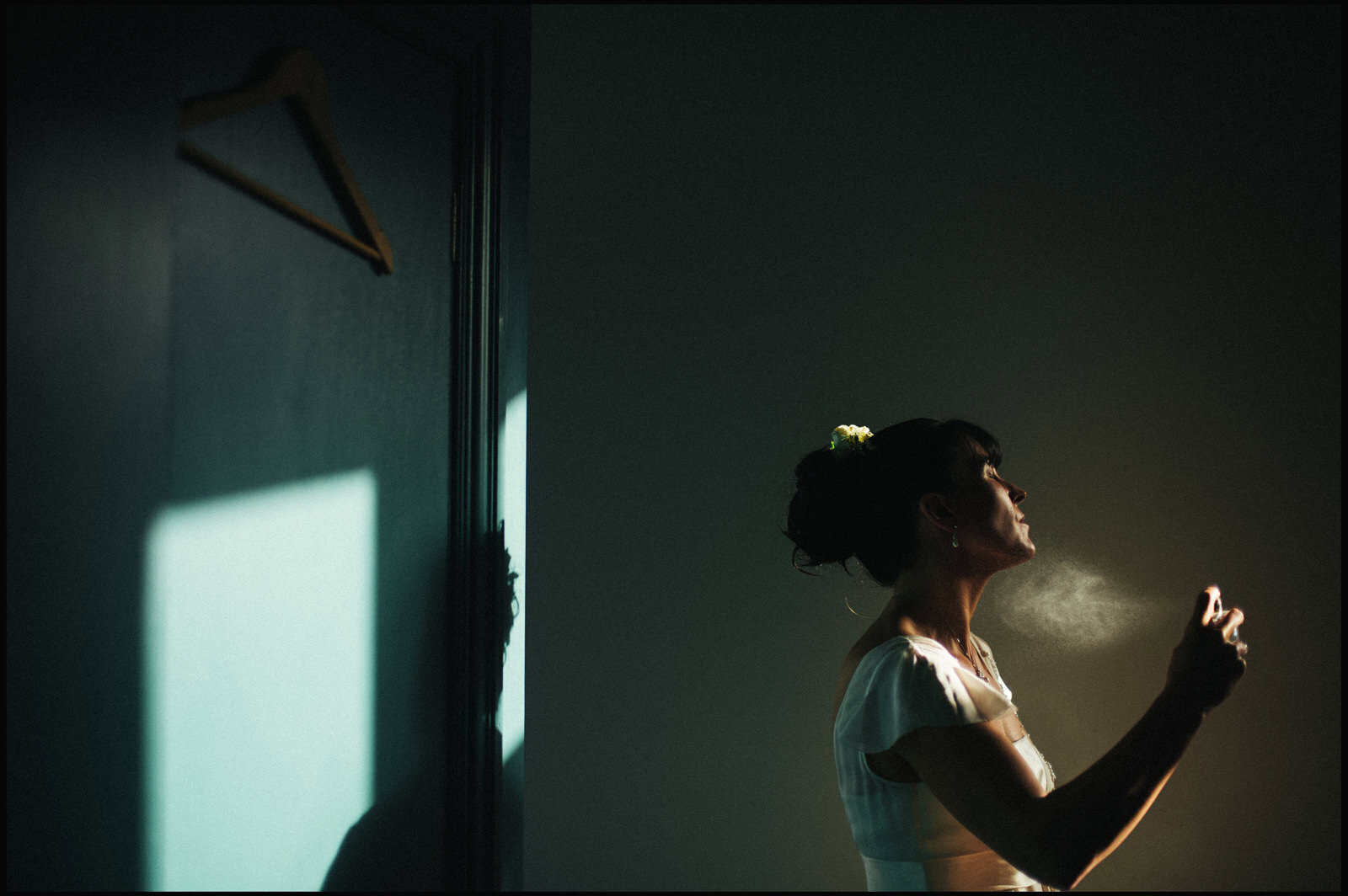 Woman in white dress standing in a dimly lit room, with sunlight casting shadows on the wall behind her, holding a spray bottle near her face.