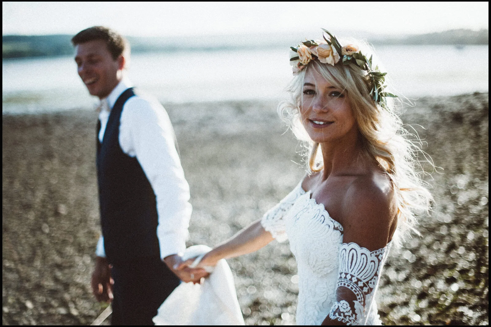 A bride and groom holding hands on a beach, with the bride in a lace off-shoulder wedding dress and a flower crown, smiling and looking at the camera, and the groom in a white shirt and black vest, smiling and looking away.