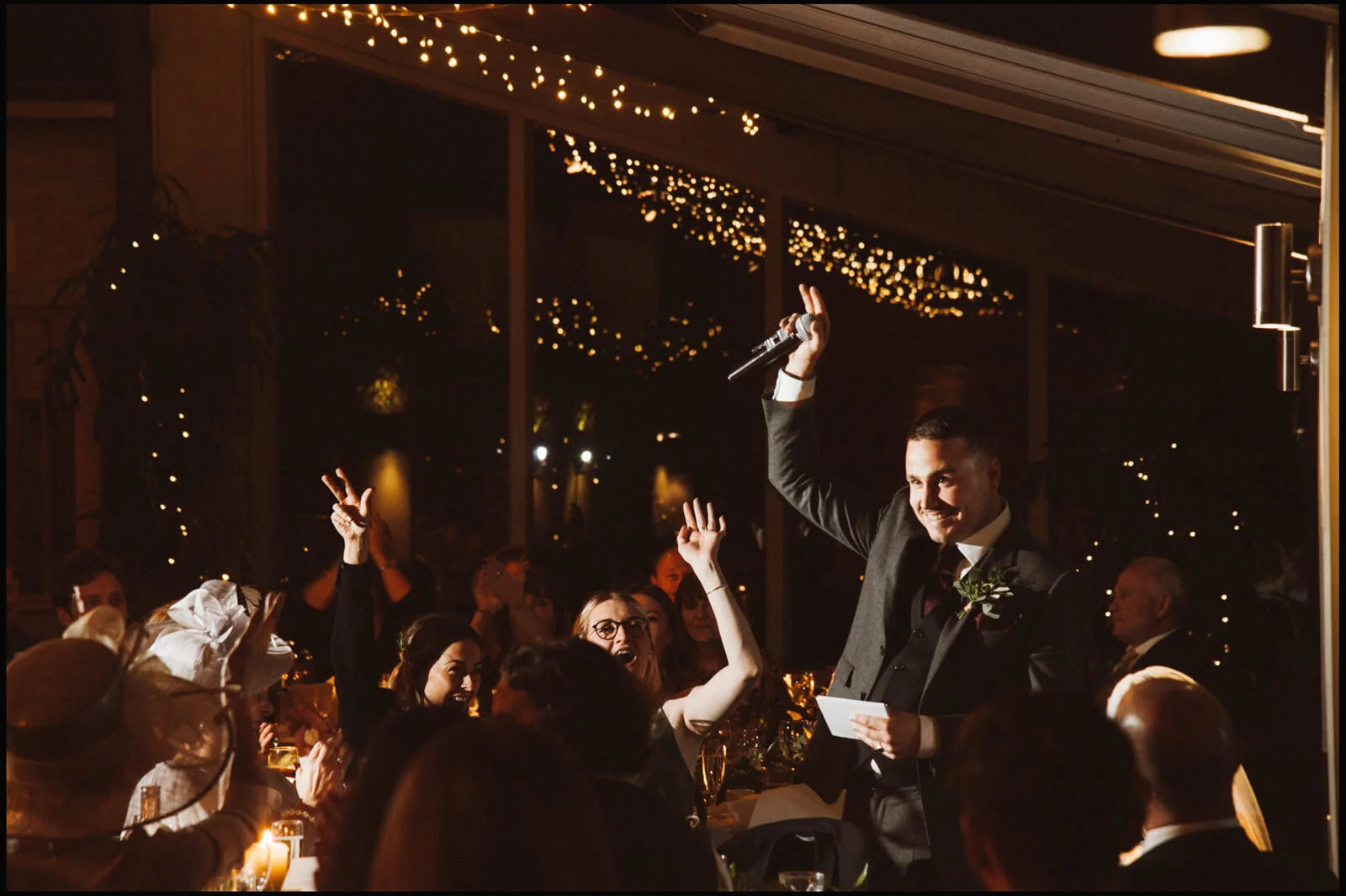 A man in a suit holding a microphone and a piece of paper, smiling and raising his hand in front of a group of people at an indoor event with string lights and festive decorations.