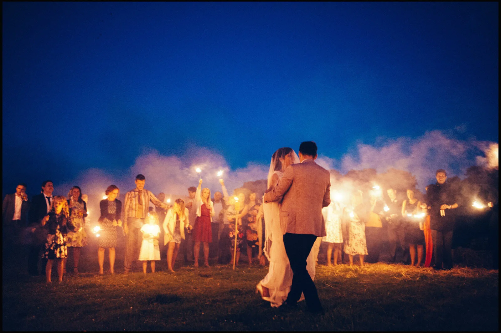 A bride and groom dancing outdoors at night surrounded by people holding sparklers.