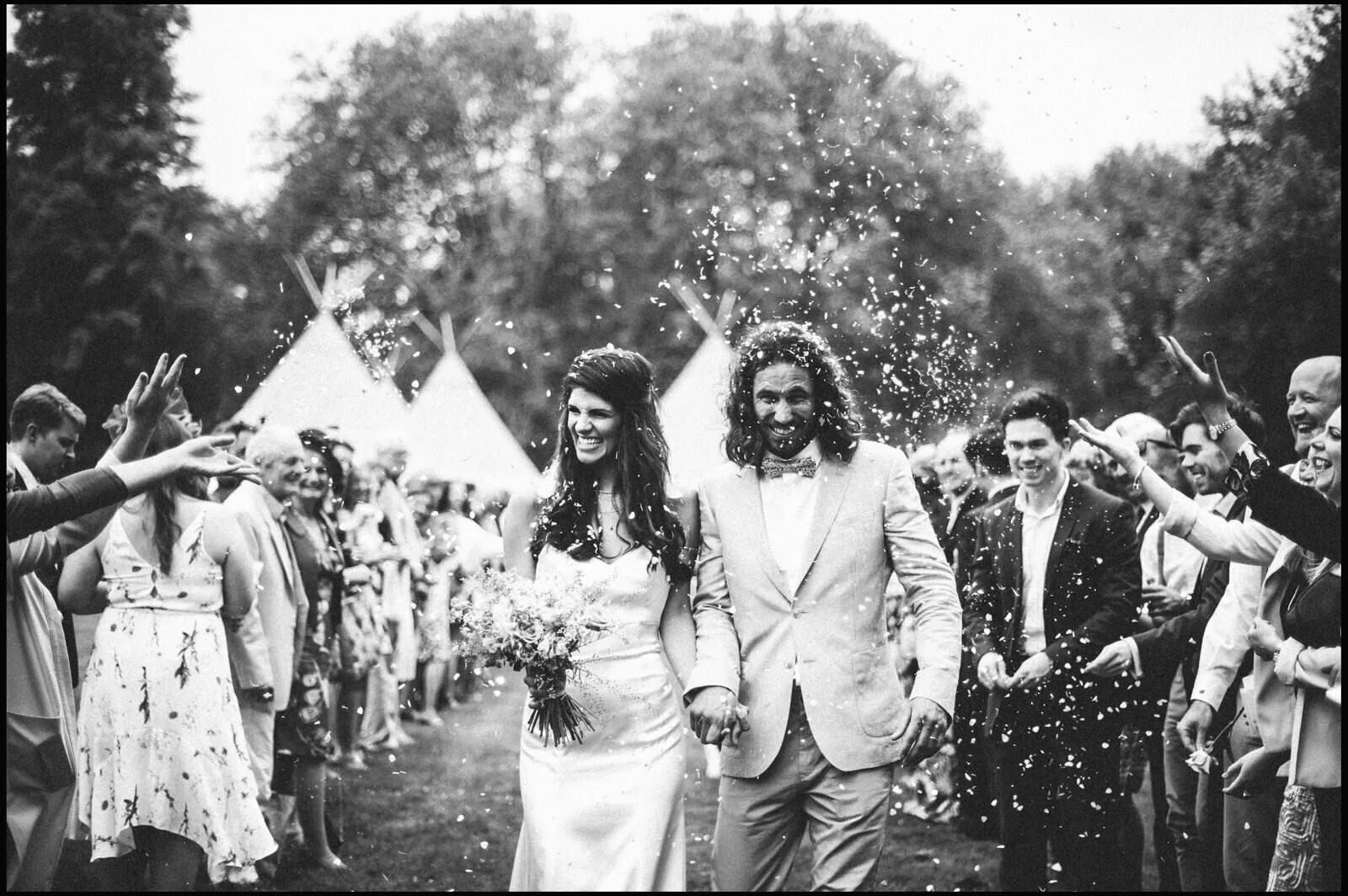 Black and white photo of a bride and groom walking hand in hand through a crowd of wedding guests, surrounded by confetti, outdoors with trees and tents in the background.