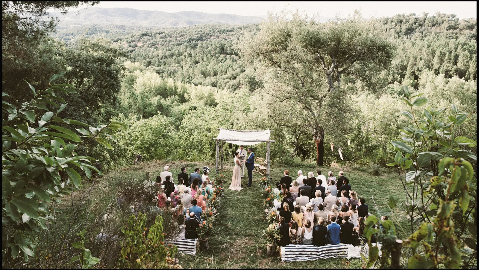 Outdoor wedding ceremony on a grassy clearing surrounded by trees and mountains, with bride and groom standing under a small wooden arch, guests seated on benches and chairs watching the ceremony.
