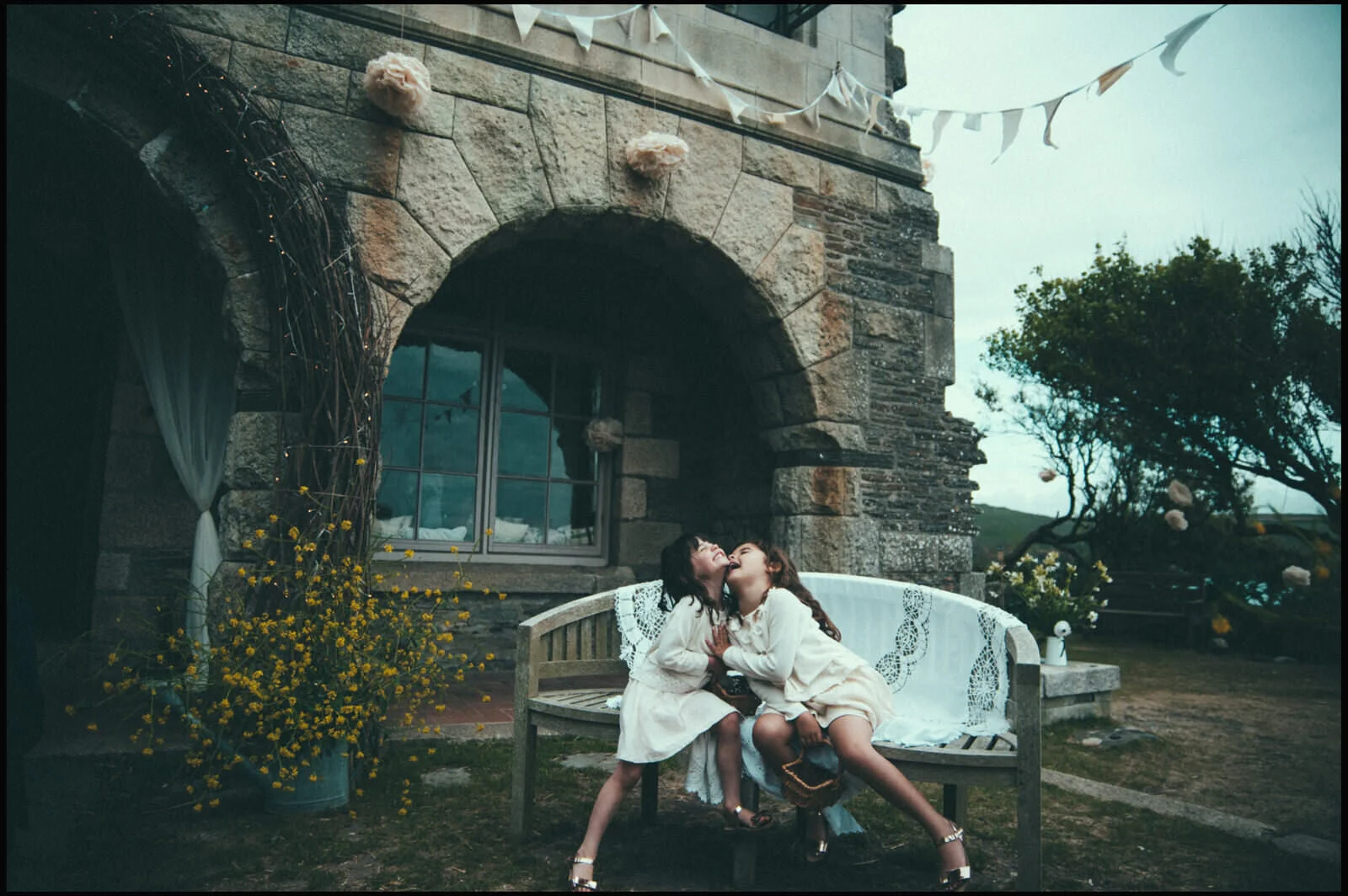 Two women in white dresses sitting close on a wooden bench outside a stone building, sharing a moment of joy, with flowers and outdoor decorations around.