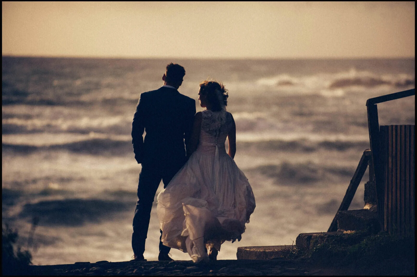 A couple dressed in formal attire walking along the beach at sunset, with the ocean and waves in the background.