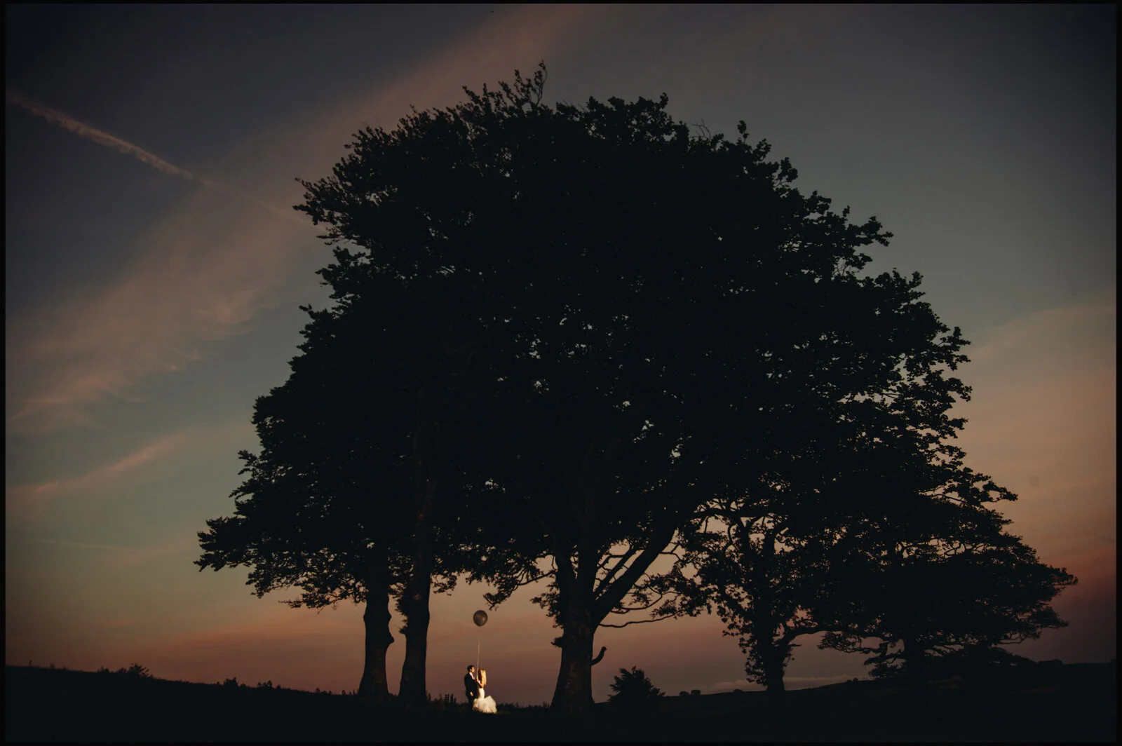 Silhouetted wedding couple standing under large trees at sunset with a balloon.