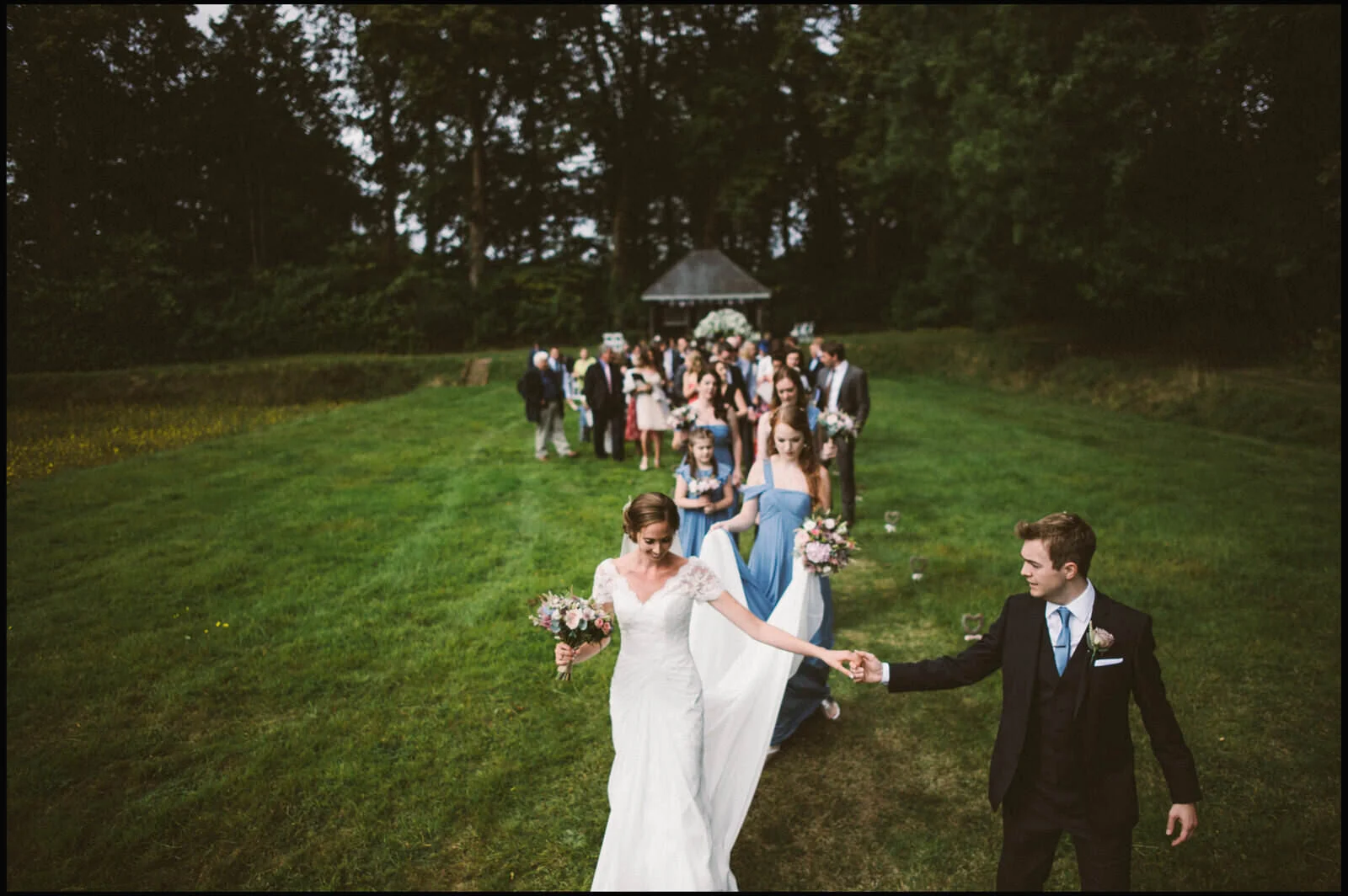 A bride and groom holding hands and walking on a grassy field surrounded by wedding guests at an outdoor wedding ceremony, with trees in the background.