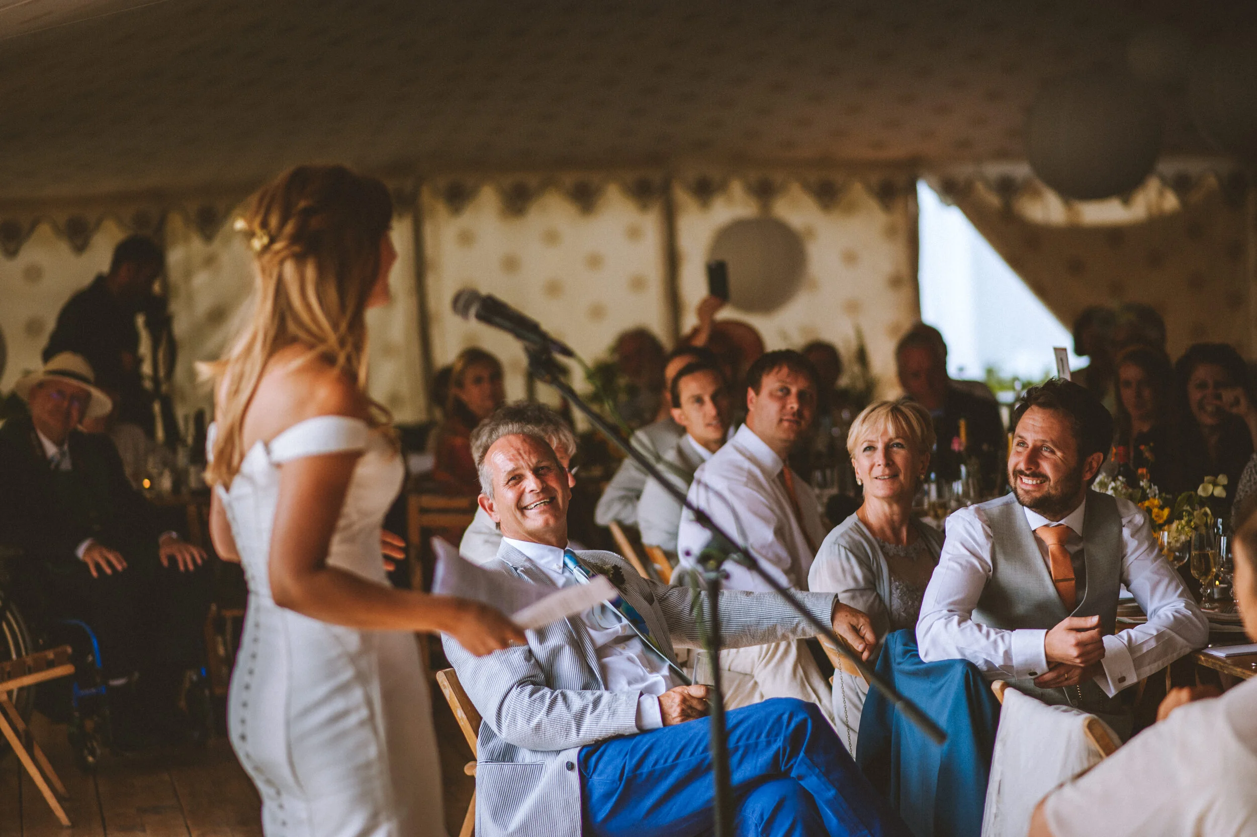 A woman in a white off-shoulder dress giving a speech at a wedding reception, standing before seated guests who are smiling and listening, in a decorated tent with floral arrangements.