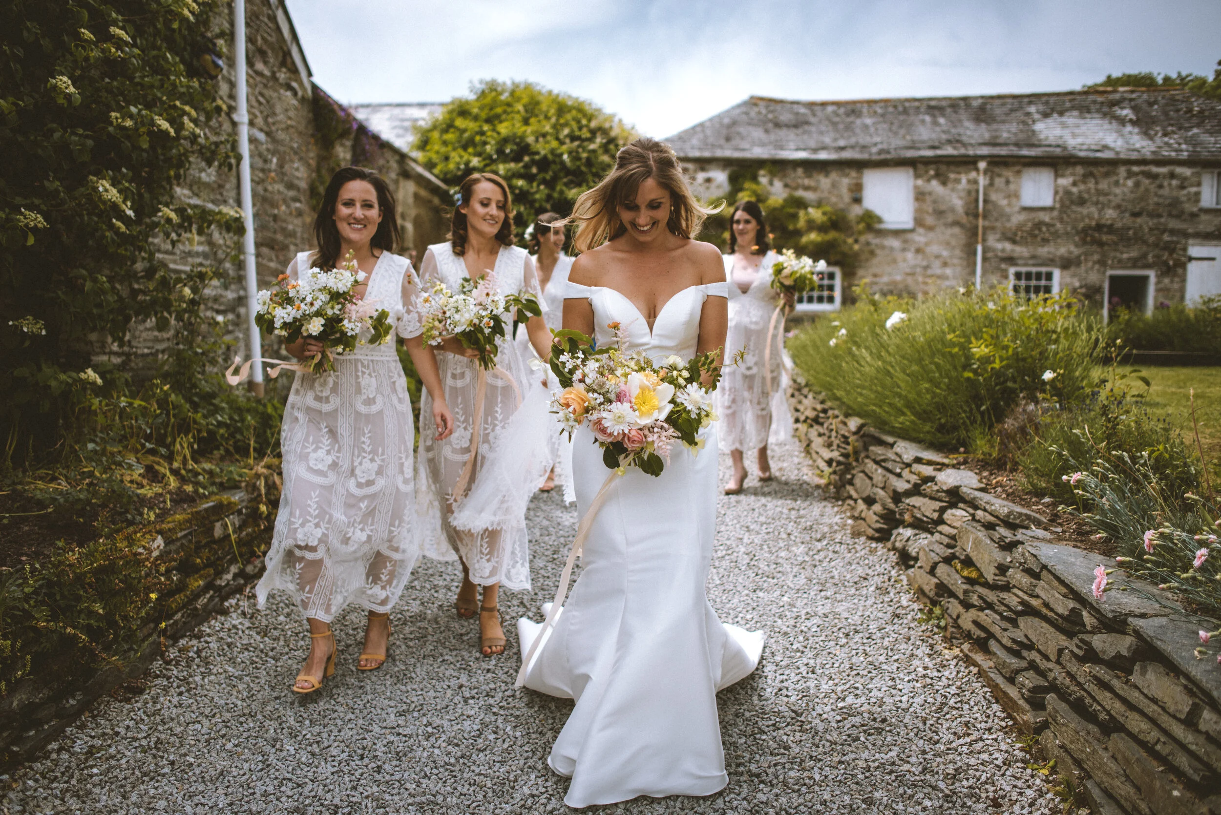 Bride walking with her bridesmaids outdoors, all holding bouquets, in a garden setting with stone buildings and greenery.