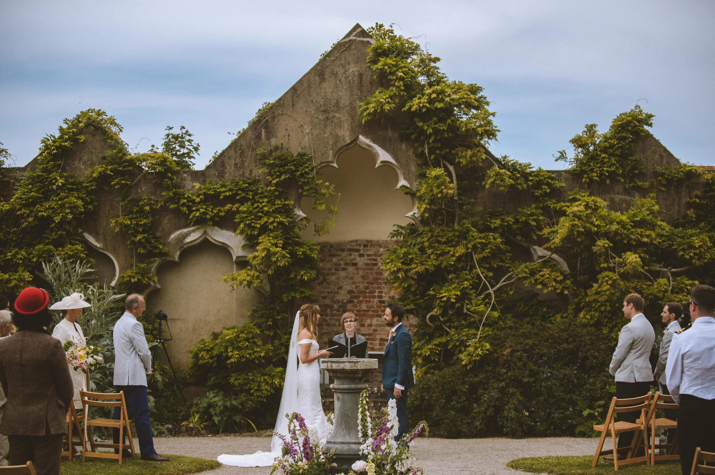 A wedding ceremony outdoors in front of a stone wall covered with green vines, with the bride and groom standing at a small altar, surrounded by guests.