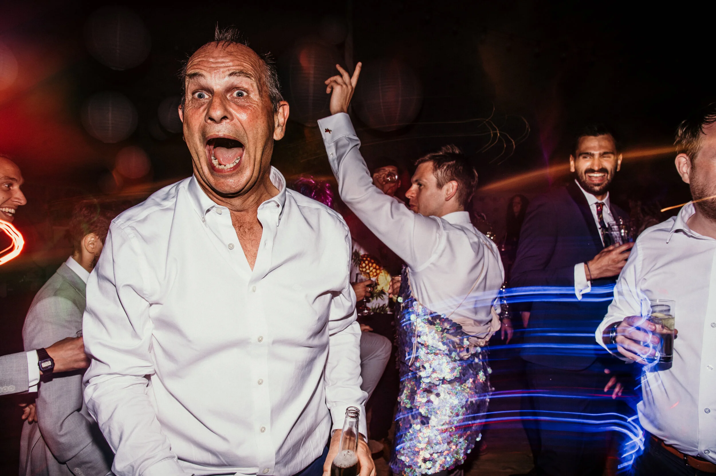 A lively party scene with people dancing and having fun, including a man in the foreground with an exaggerated expression, opening his mouth wide and holding a bottle, while others in the background are smiling and dancing.