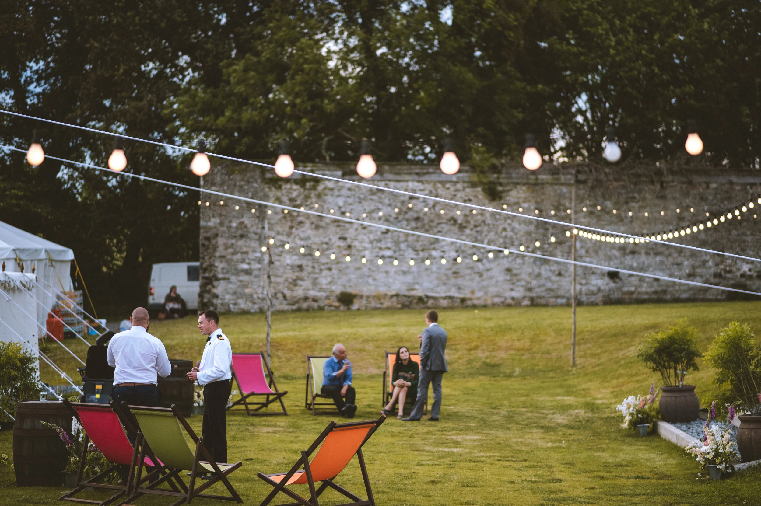 People socializing outdoors at evening with string lights, lawn chairs, and a stone wall in the background.