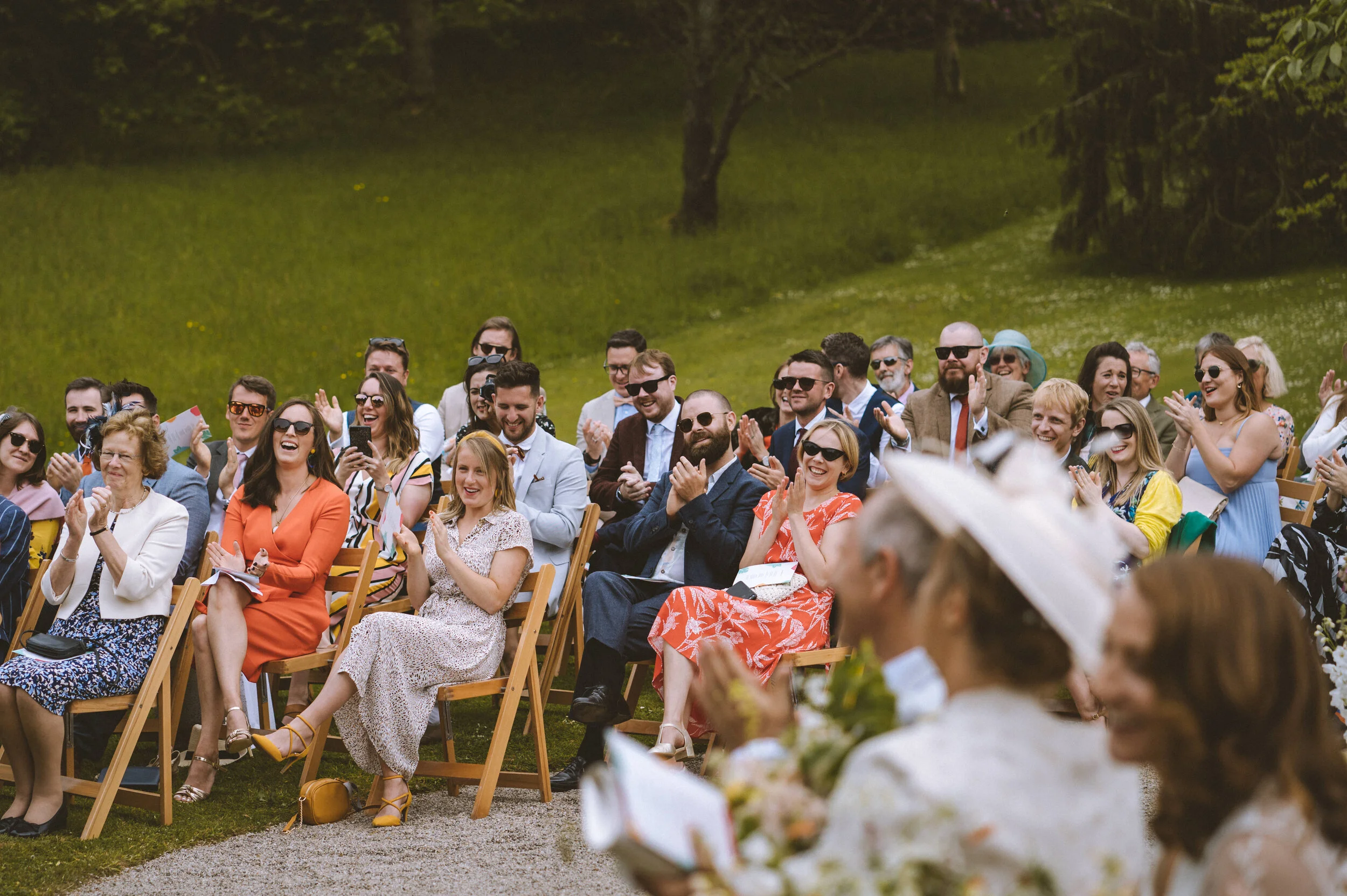 A group of people at an outdoor wedding ceremony, seated on wooden chairs on a grassy area, clapping and smiling.