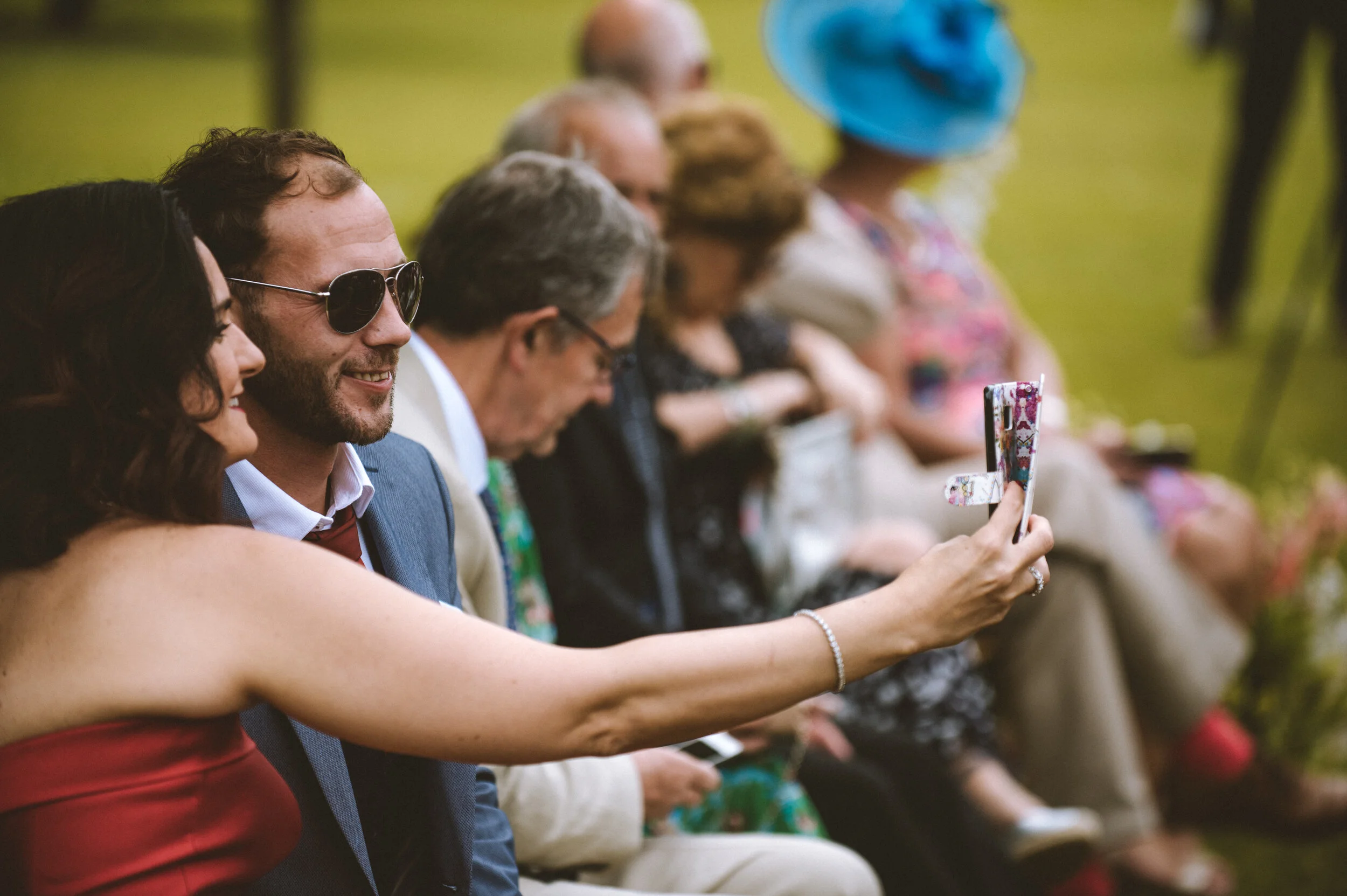 People sitting outdoors, with some dressed formally, others casual, during a social event or wedding ceremony. A woman with a bracelet is taking a photo of a smiling man wearing sunglasses.