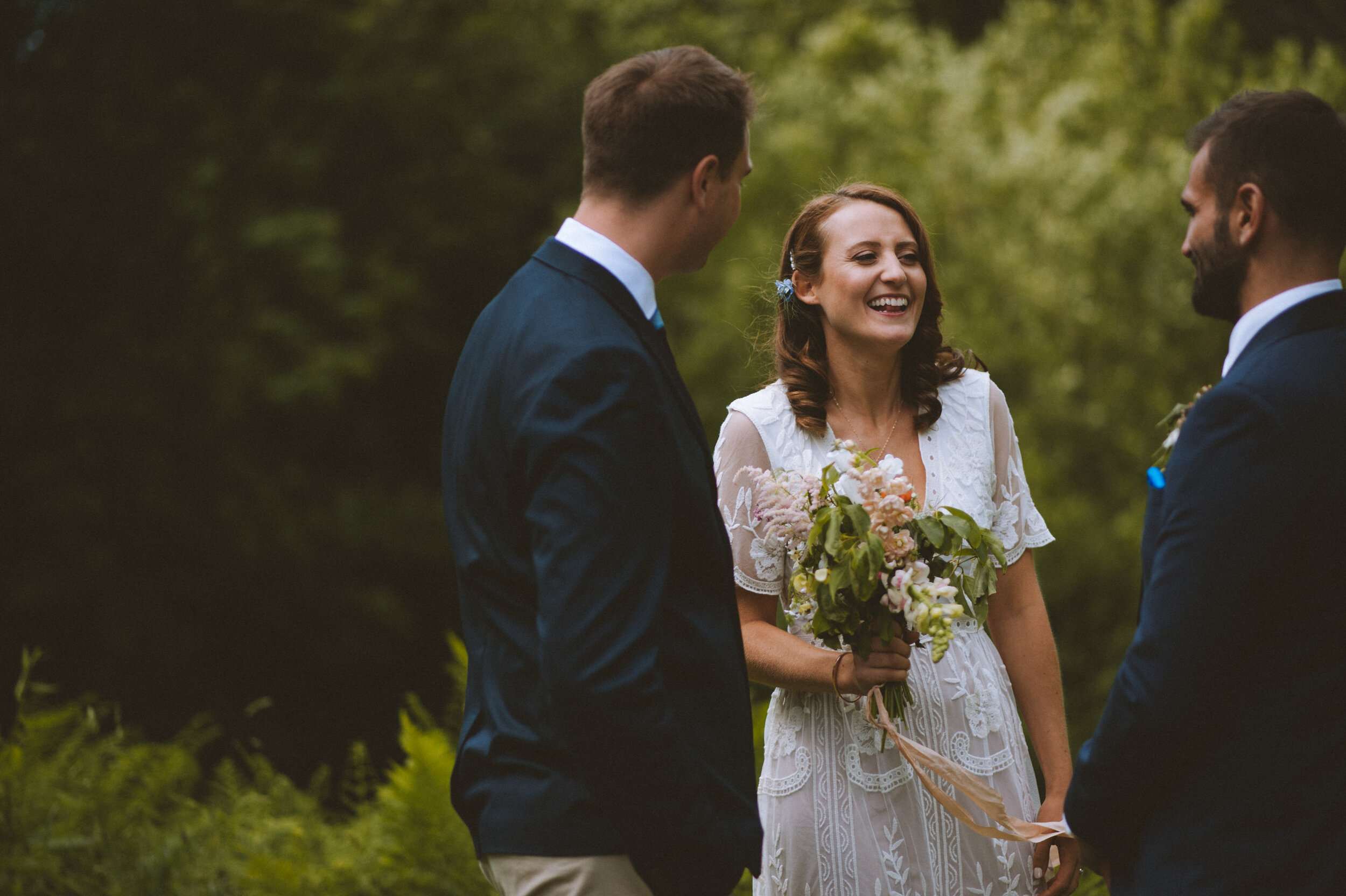 A woman in a white lace dress holding a bouquet of flowers smiling at two men in suits during an outdoor wedding ceremony.