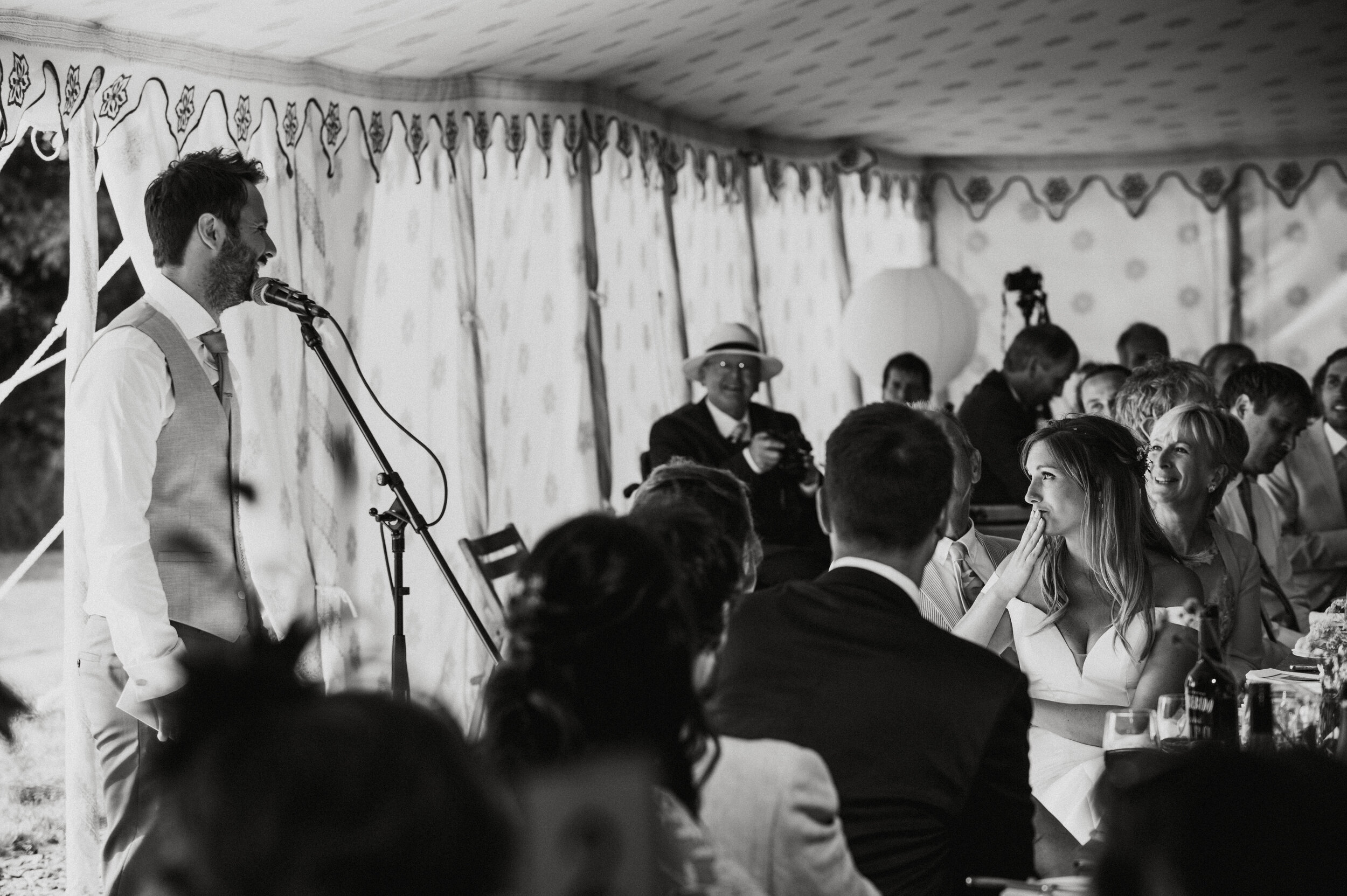A man is giving a speech at a wedding reception under a decorated tent, with guests seated and listening, some smiling and interacting.