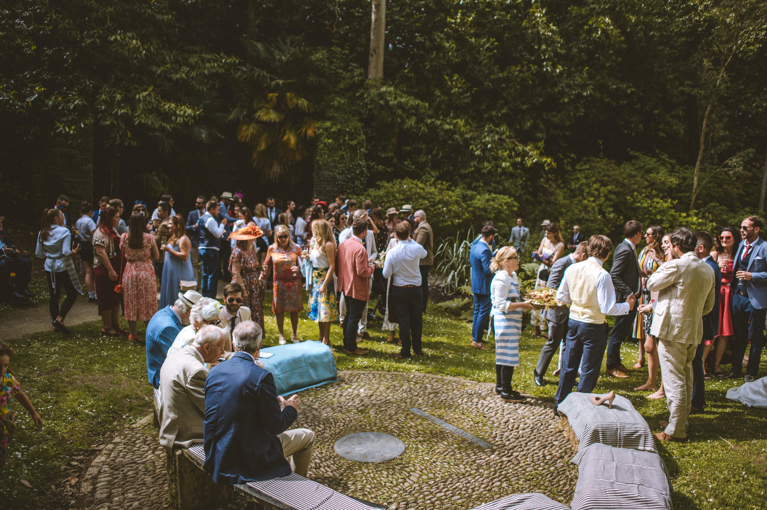 Pencarrow House wedding photography in Cornwall - People gathered outdoors for a social event or celebration, standing on a lawn surrounded by trees and greenery.