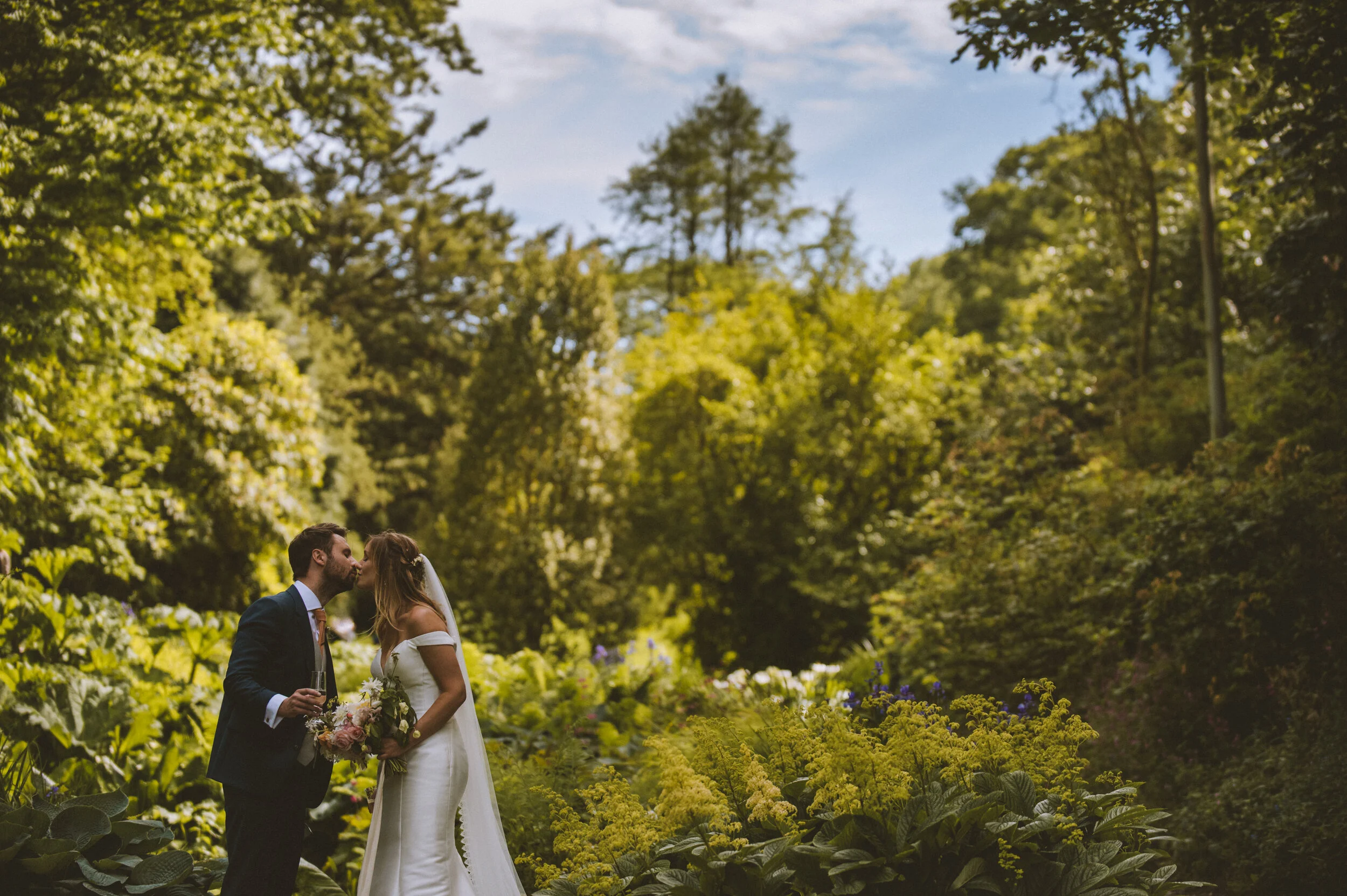 Pencarrow House wedding photography in Cornwall - A bride and groom kissing outdoors in a lush green forest, with the groom holding a glass of champagne and the bride holding a bouquet of flowers.