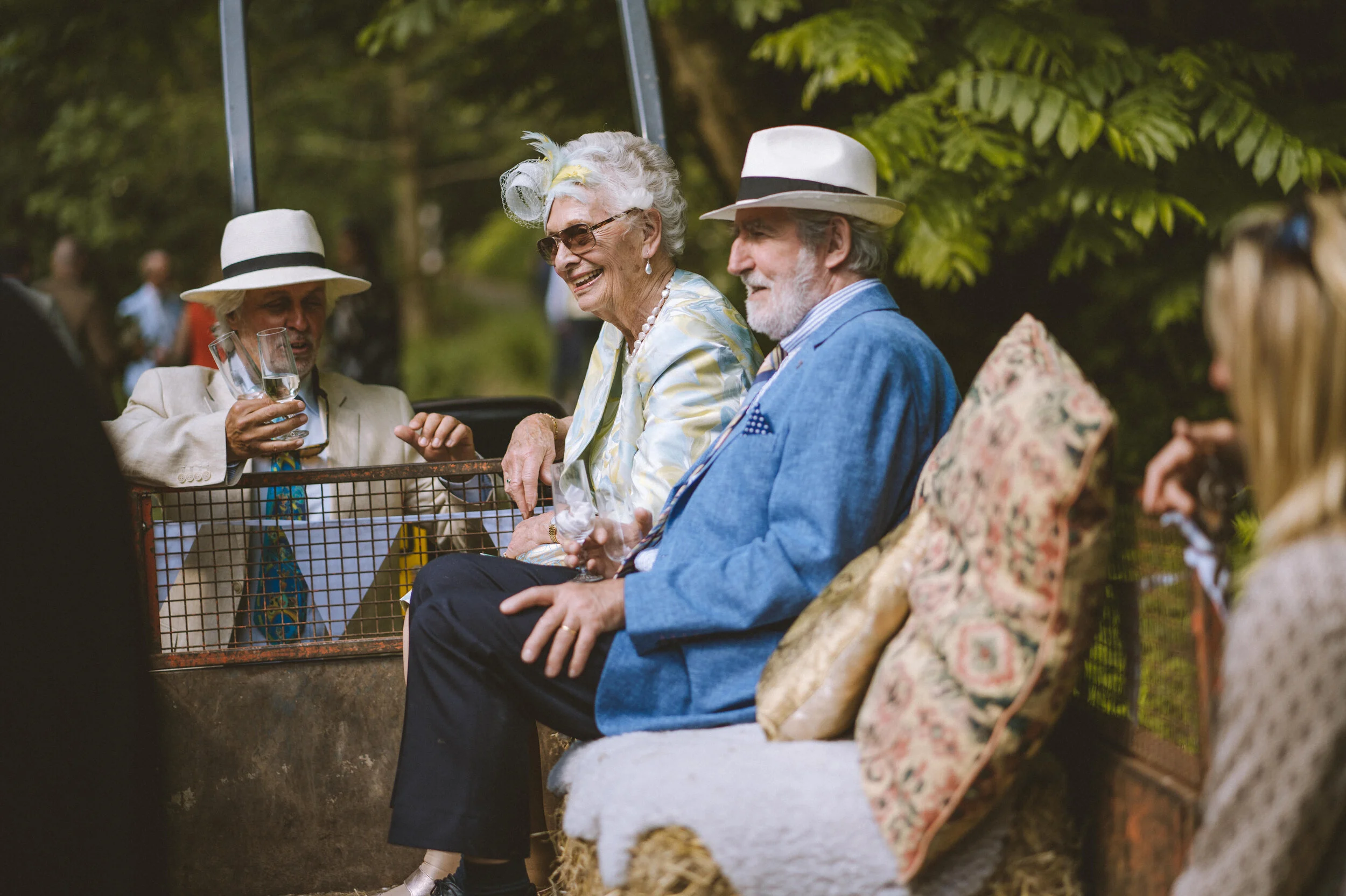 Four elderly people sitting outdoors on a bench and in a cart, dressed in vintage clothing, enjoying drinks and conversation among greenery.