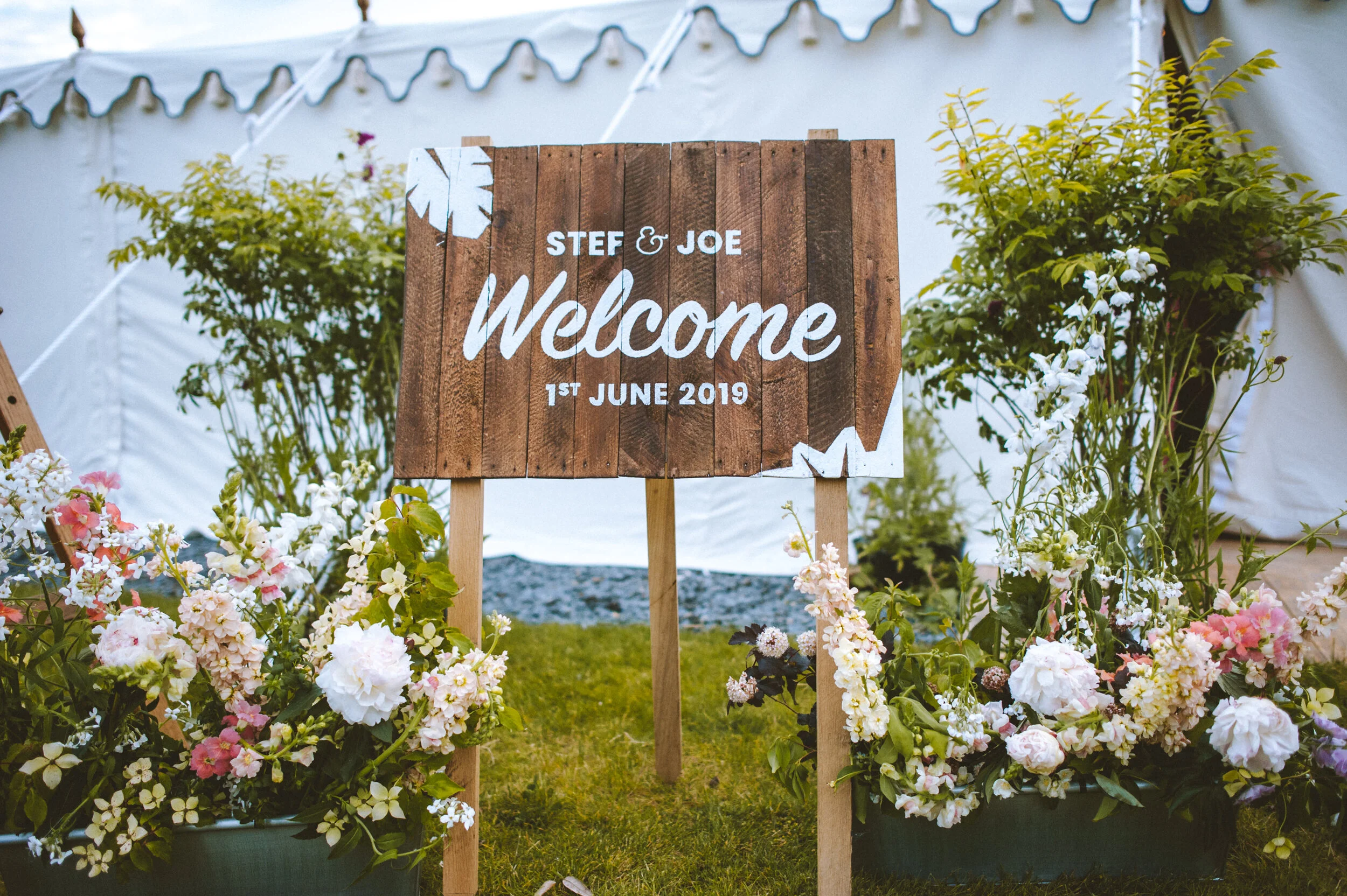 Wooden welcome sign for Stef & Joe dated June 1, 2019, surrounded by colorful flowers and greenery, with a white tent in the background.