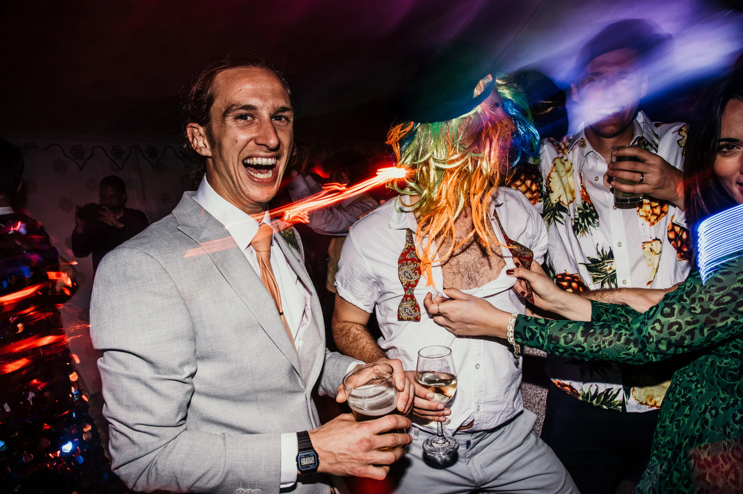 People at a party, one man in a light gray suit holding drinks, others in colorful shirts, with party hats, in a lively celebration with vibrant lighting.