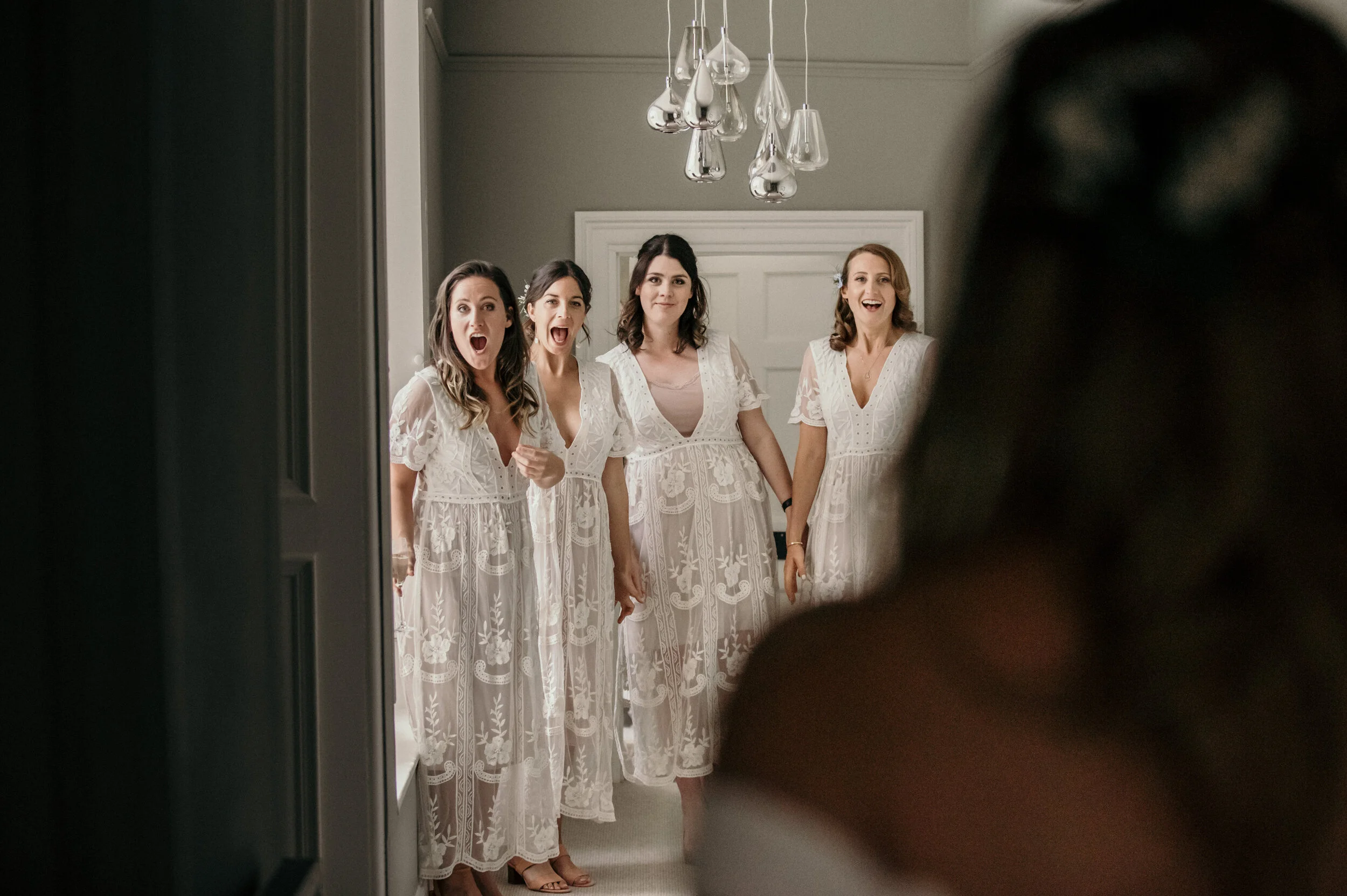 Four women in white lace dresses standing together indoors, surprised and smiling, as another woman is partially visible in the foreground.