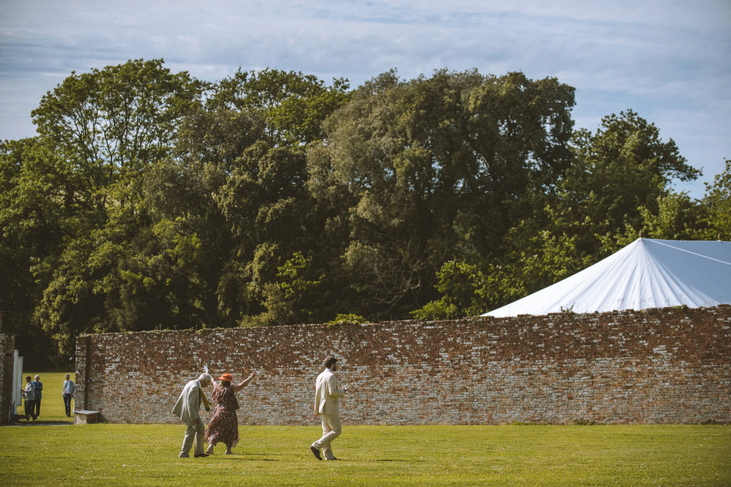 People walking on a grassy area in front of an old brick wall with a white tent behind it, surrounded by green trees and a cloudy sky.