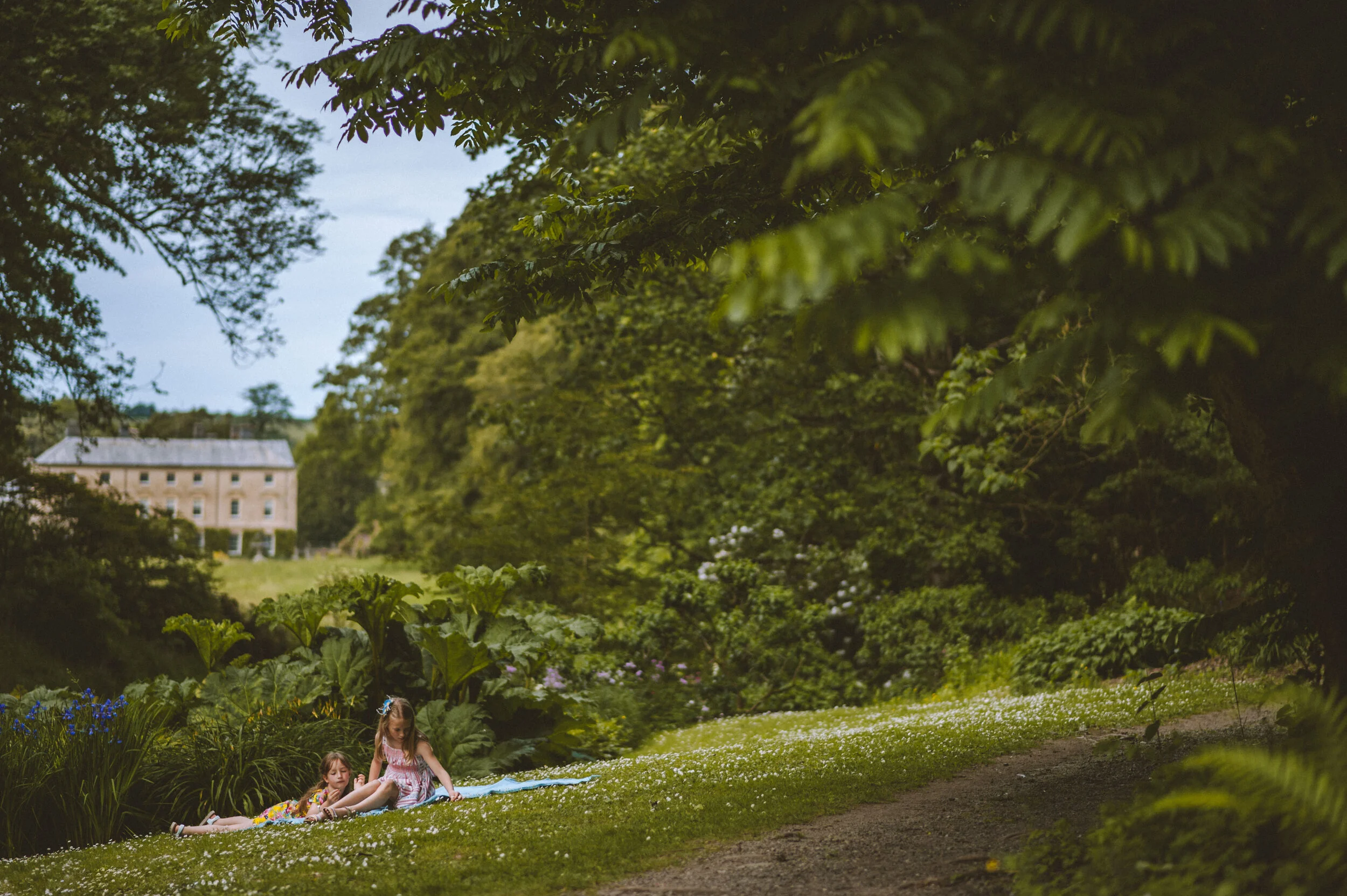 Two young girls are sitting on a blanket on a grassy area in a lush garden, surrounded by tall trees and green foliage, with a large building visible in the background.