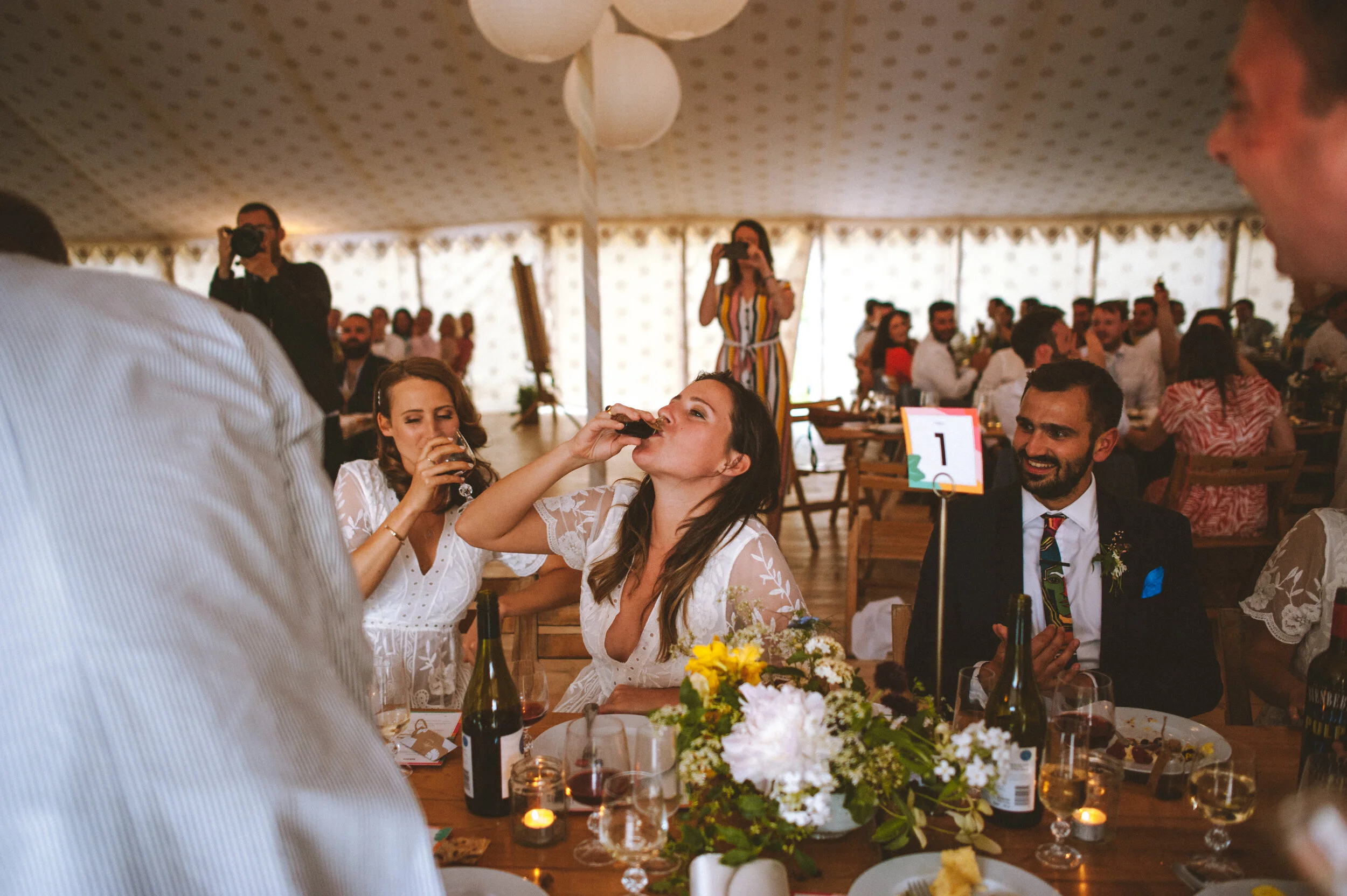 People seated at a table during a wedding reception, with two women drinking and smiling, a man in a suit, floral centerpiece, and guests in the background under a decorated tent.