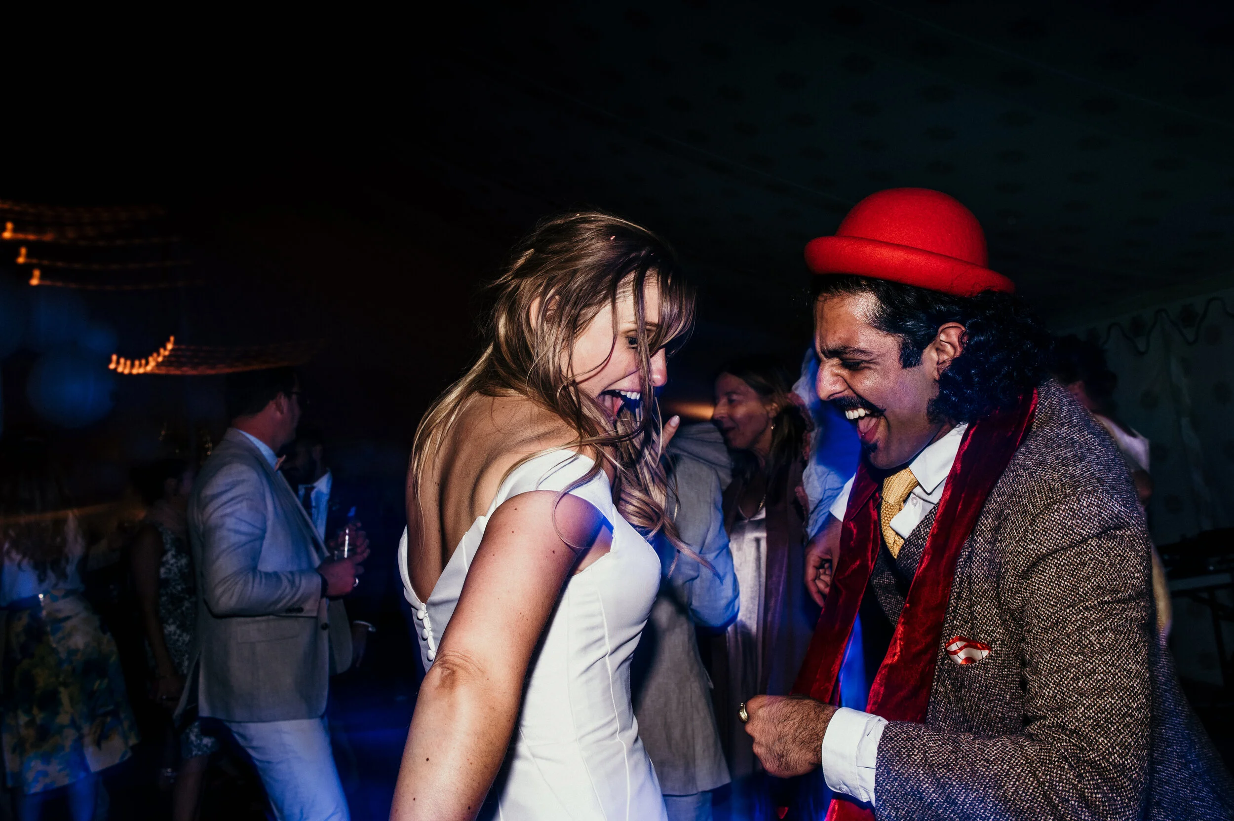 People dancing and laughing at a party, with a woman in a white dress and a man in a red hat in the foreground.
