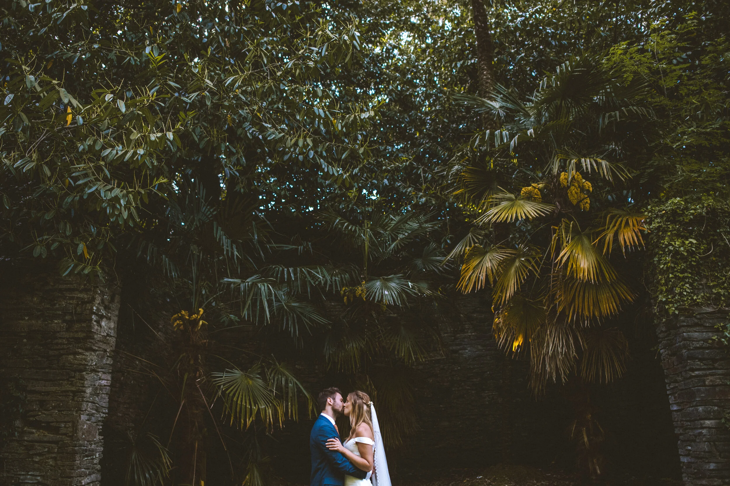 A couple in wedding attire sharing a kiss, surrounded by lush green tropical foliage and trees.