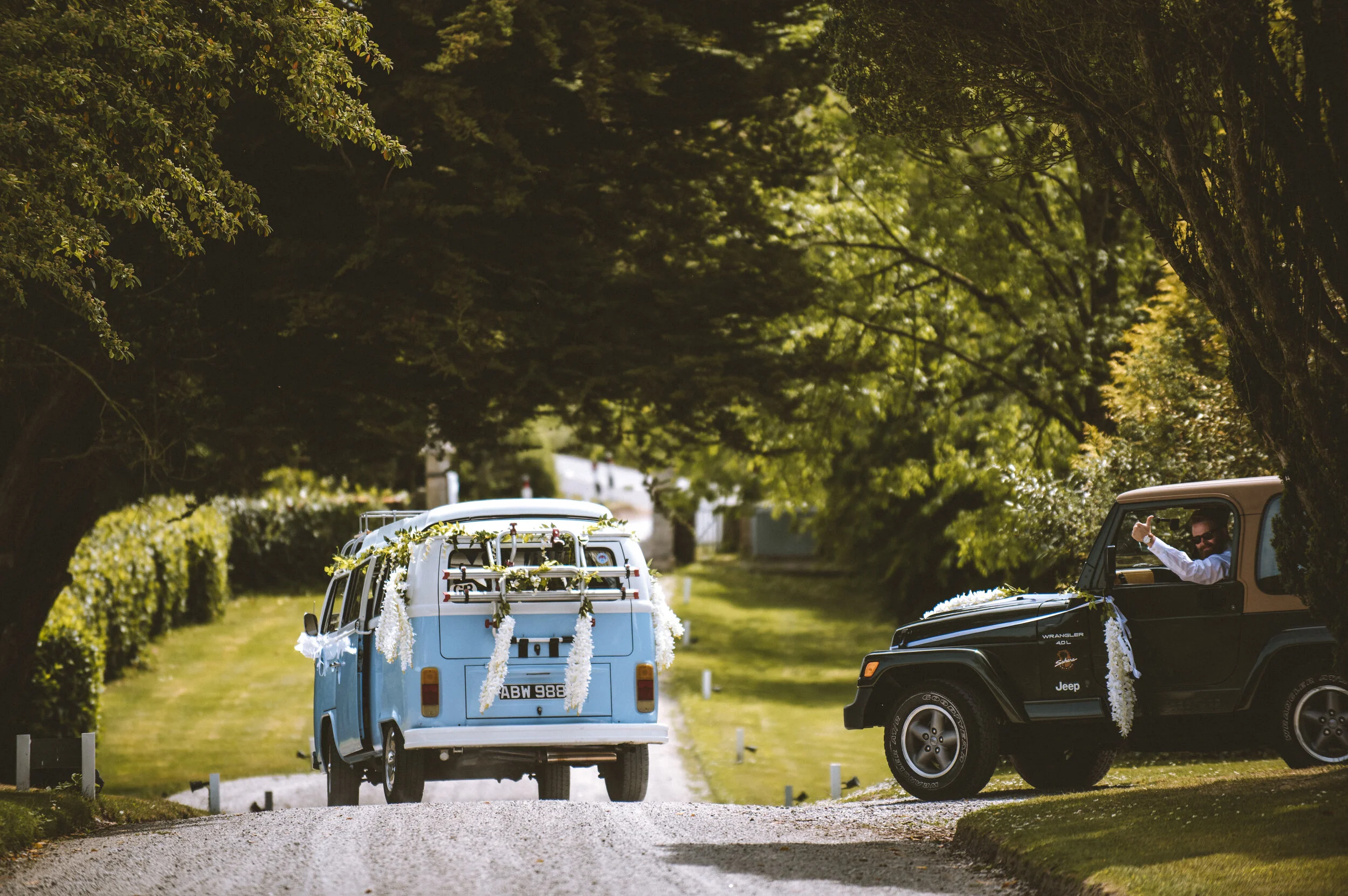 A vintage blue van and a black jeep decorated with flowers, parked under a canopy of trees on a country road, likely at a wedding or special event.