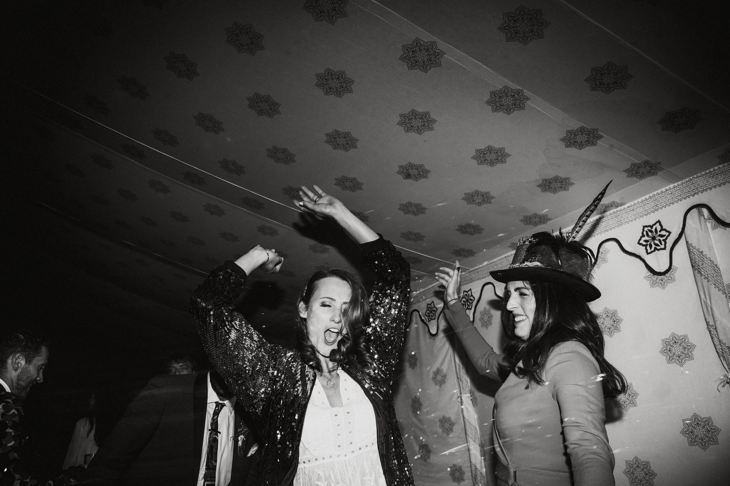 Two women dancing and celebrating at a party, one wearing a sparkly jacket and the other a hat with a feather, in a decorated room.