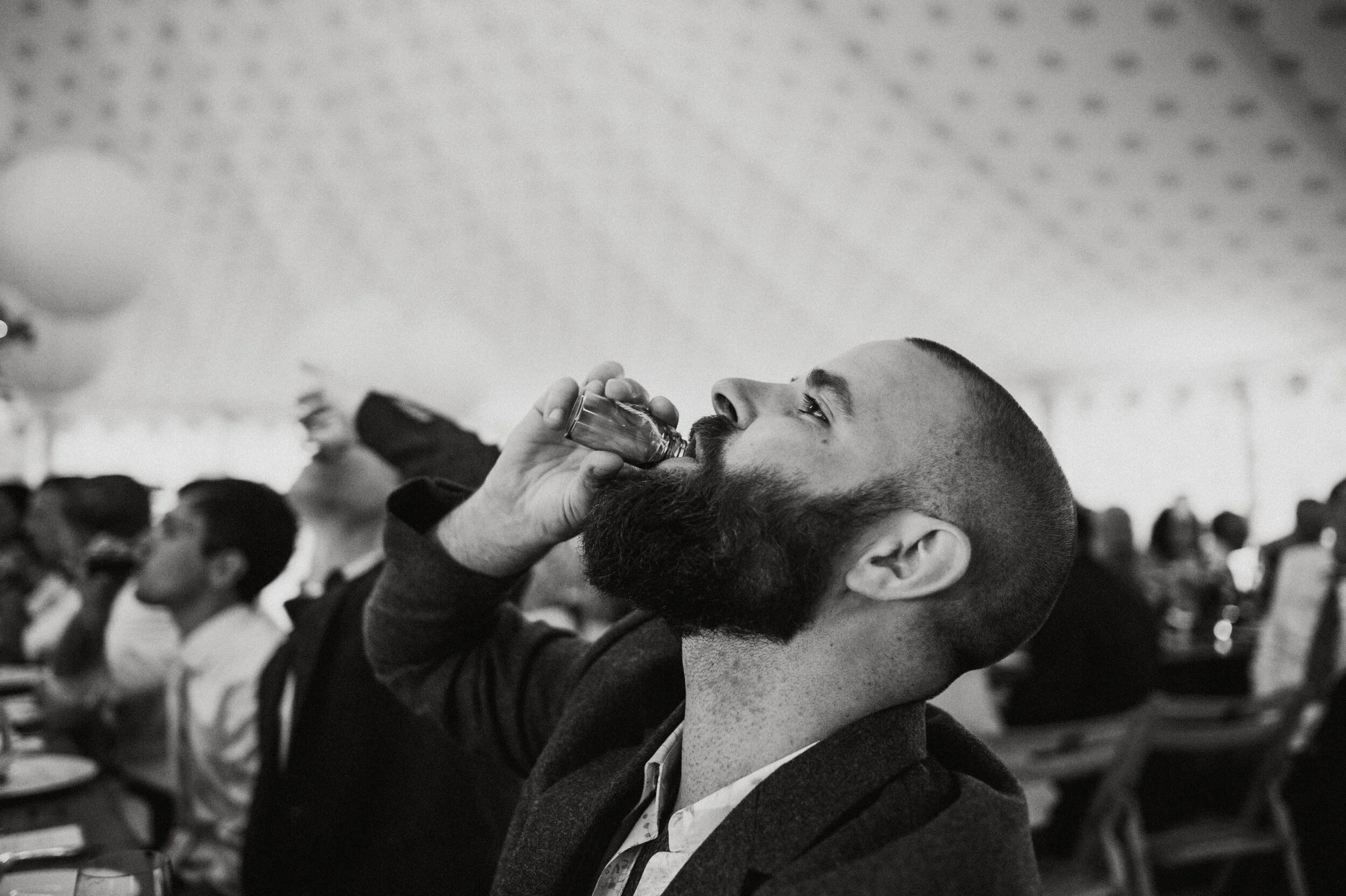 A man with a beard and short hair drinking a shot at a social gathering, with other people seated at tables in the background.