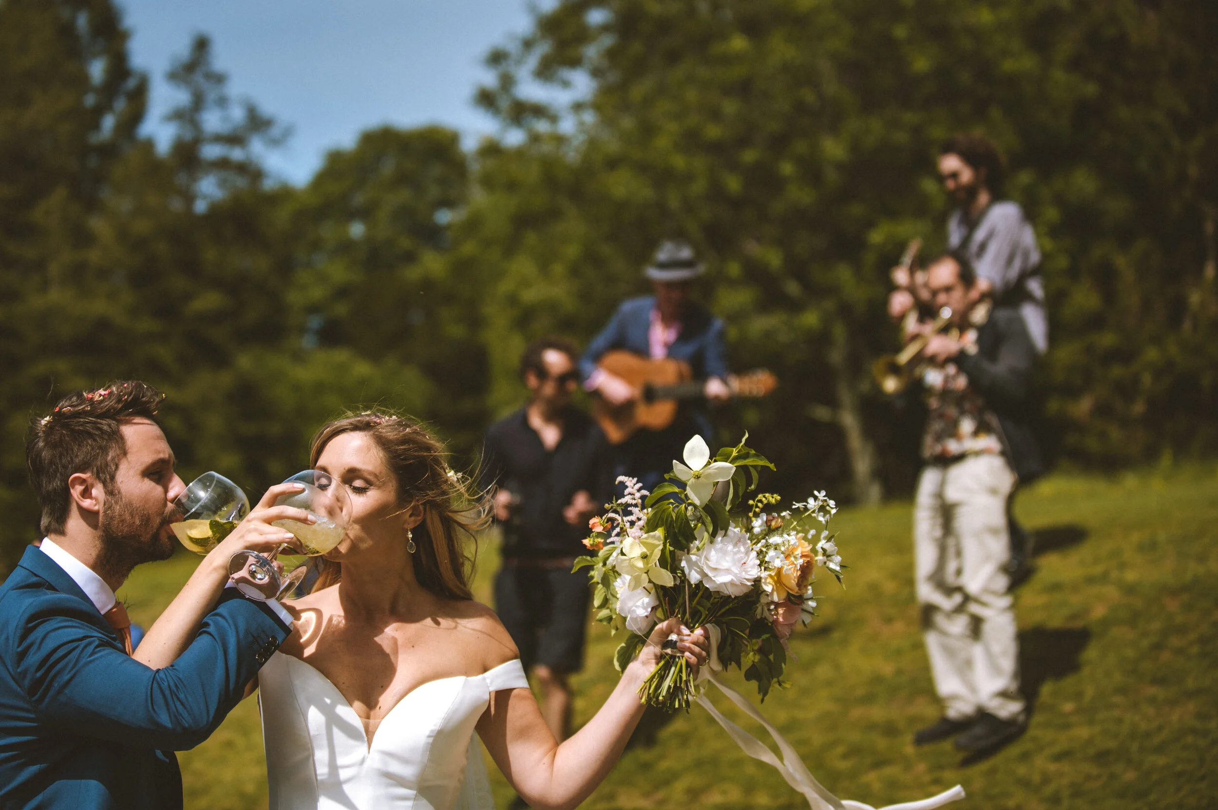 A newlywed couple toasting with glasses of white wine outdoors, with a wedding band held by the bride in one hand and a bouquet of white and peach flowers, while a band of musicians plays in the background on a grassy hill with trees.