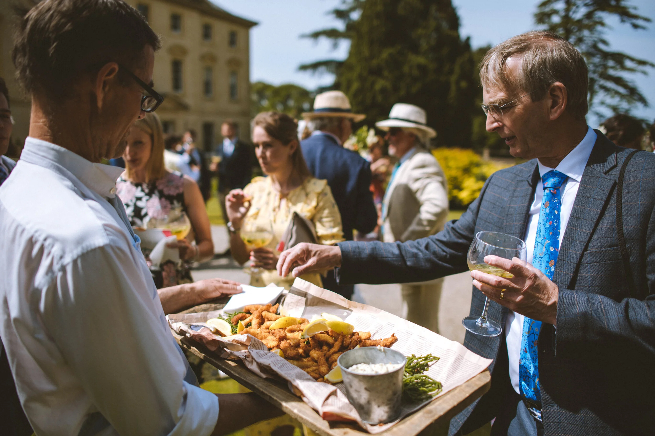 Man in a checkered suit and blue tie holding a glass of white wine, reaching for fried fish on a tray served by a waiter at an outdoor event with other guests in the background.