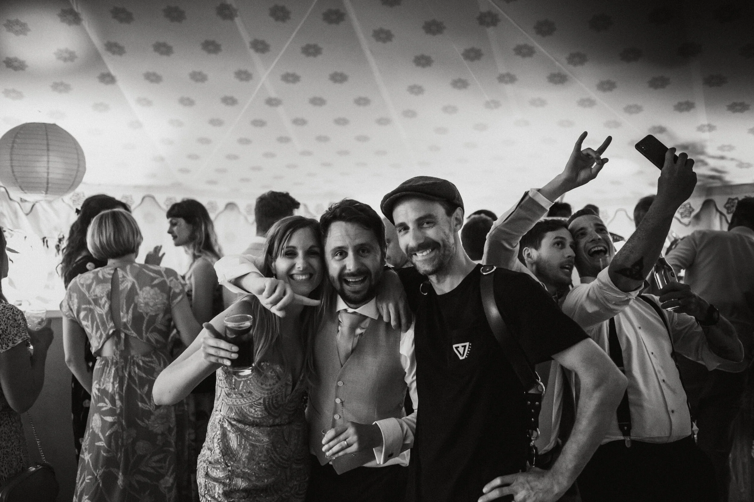 Group of people celebrating at a party, some holding drinks and taking selfies, smiling and having fun, in a decorated indoor setting.