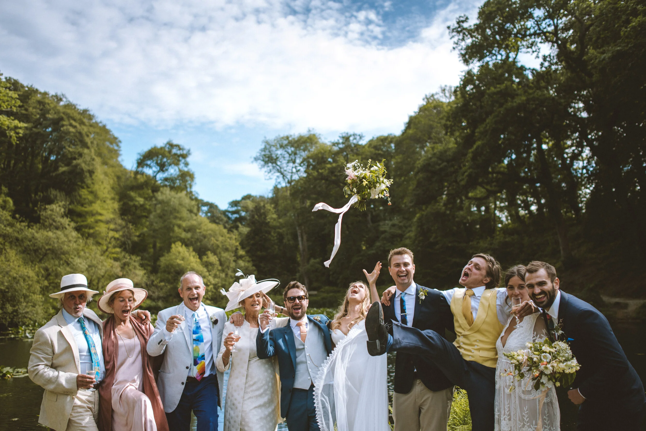 Group of people in wedding attire by a river, celebrating and throwing a bouquet into the air, surrounded by green trees under a partly cloudy sky.