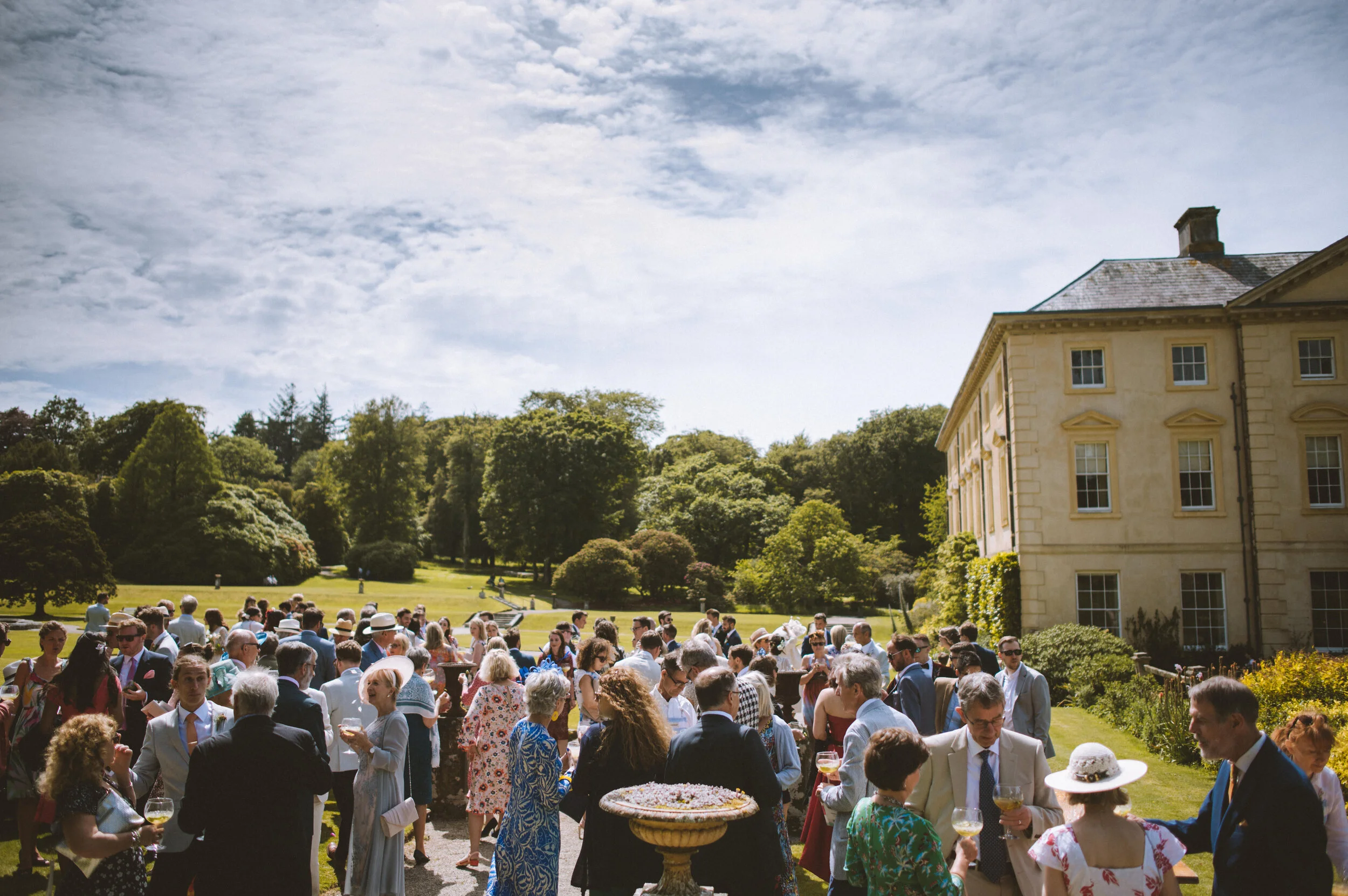 Pencarrow House wedding photography in Cornwall - Outdoor social gathering with many people dressed in formal attire, standing on a lawn next to a large, historic building, under partly cloudy skies.