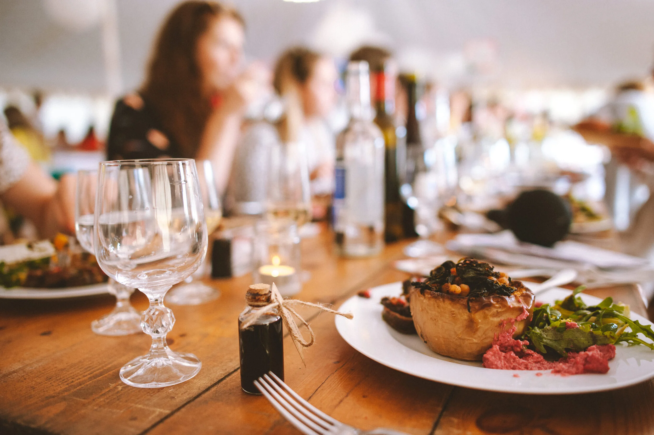 A close-up of a dinner table with empty wine glasses, a small dark bottle, and a plate containing a baked potato topped with vegetables, alongside a side salad. In the background, blurred people are seated at the table, with more bottles and a candle