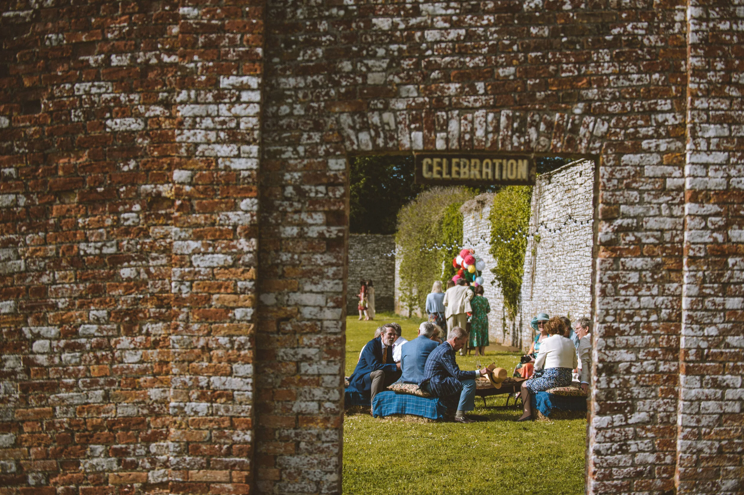 People gathered in a grassy area for a celebration event, sitting on benches and standing, with balloons and festive decorations, viewed through a brick arch.