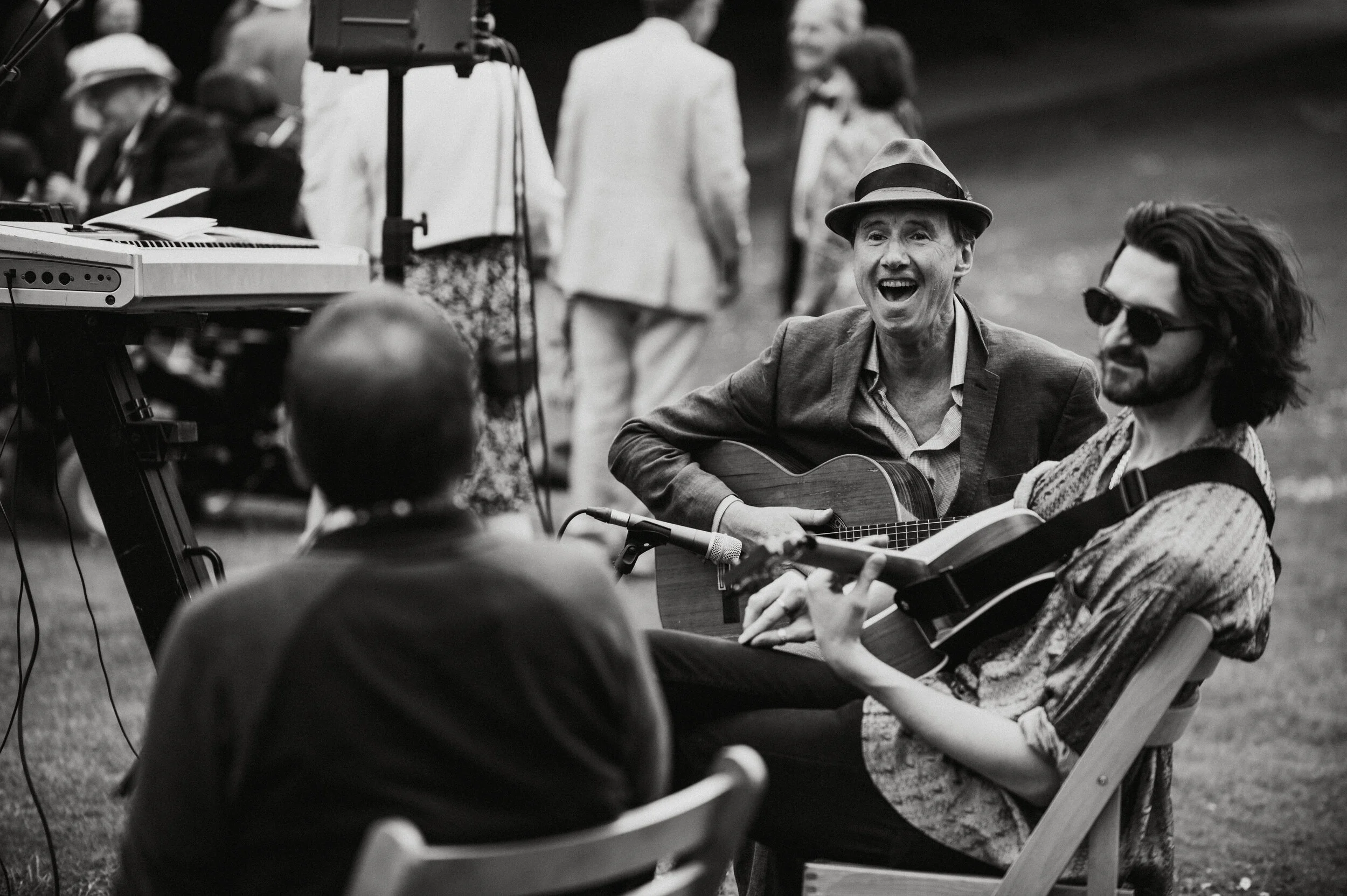 People playing guitars and singing at an outdoor gathering, with others standing and sitting nearby, some wearing sunglasses and hats.