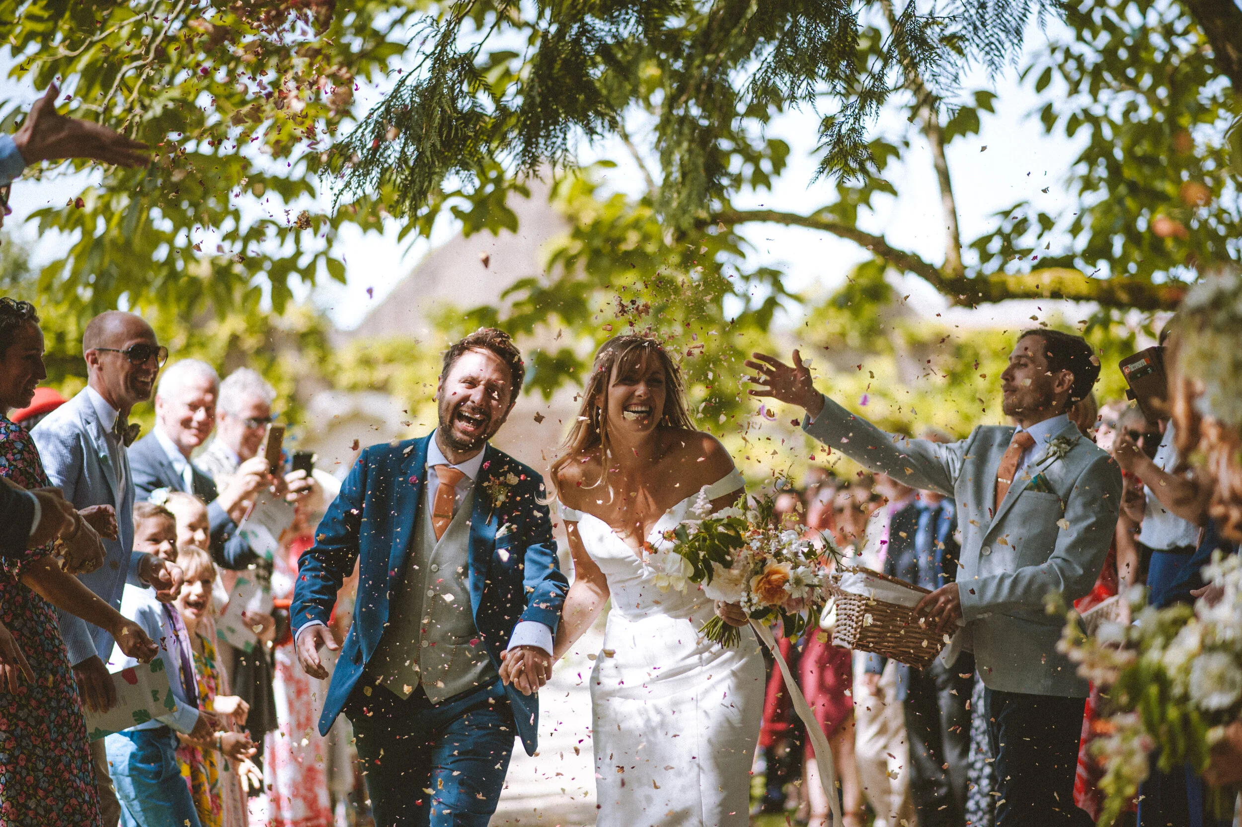 Bride and groom smiling and holding hands as they walk through a crowd of well-dressed guests during a wedding celebration, with confetti in the air and trees overhead.