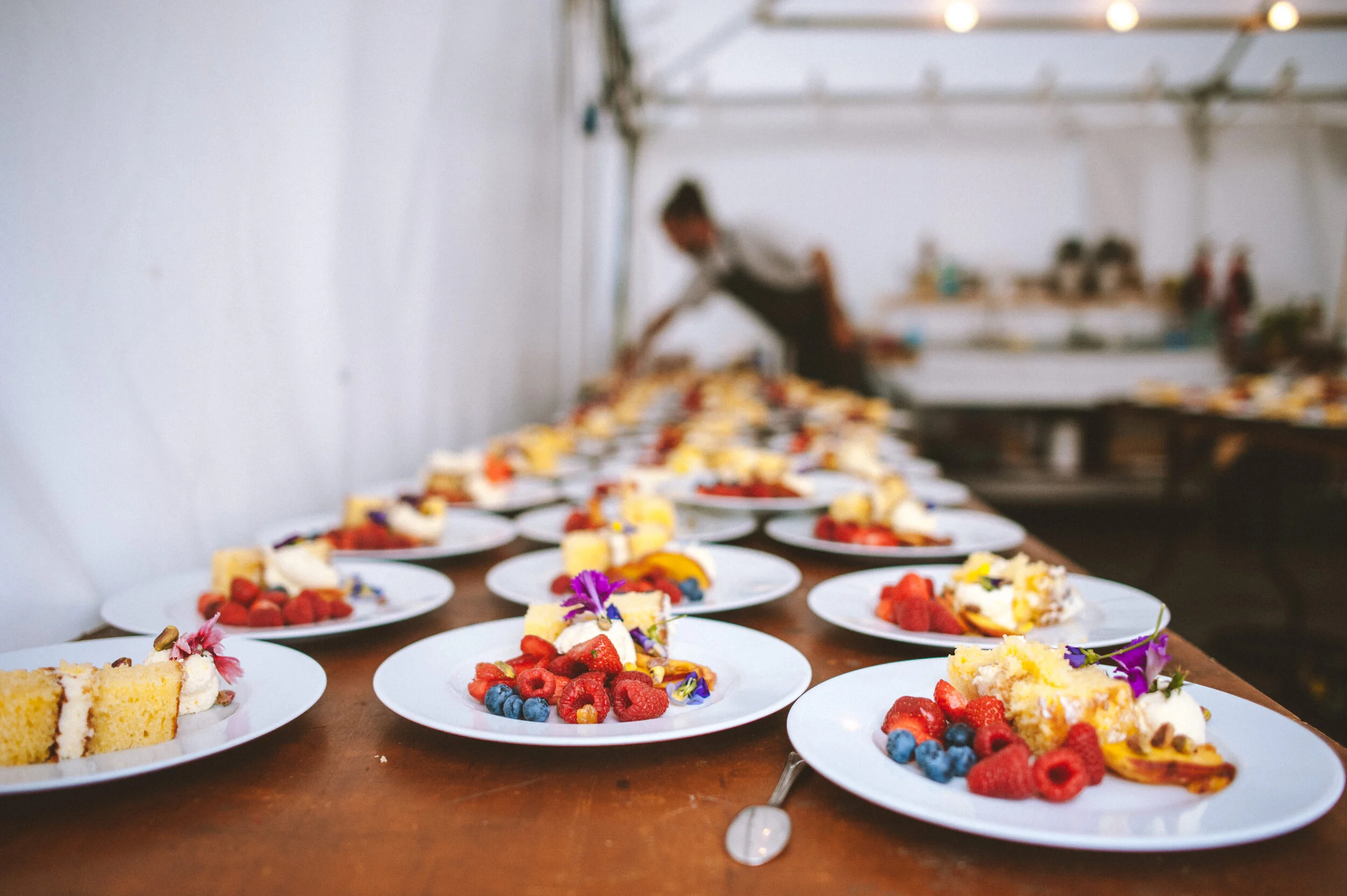 Multiple plates with slices of cake topped with fresh berries and edible flowers on a wooden table, with a blurry person arranging desserts in the background.