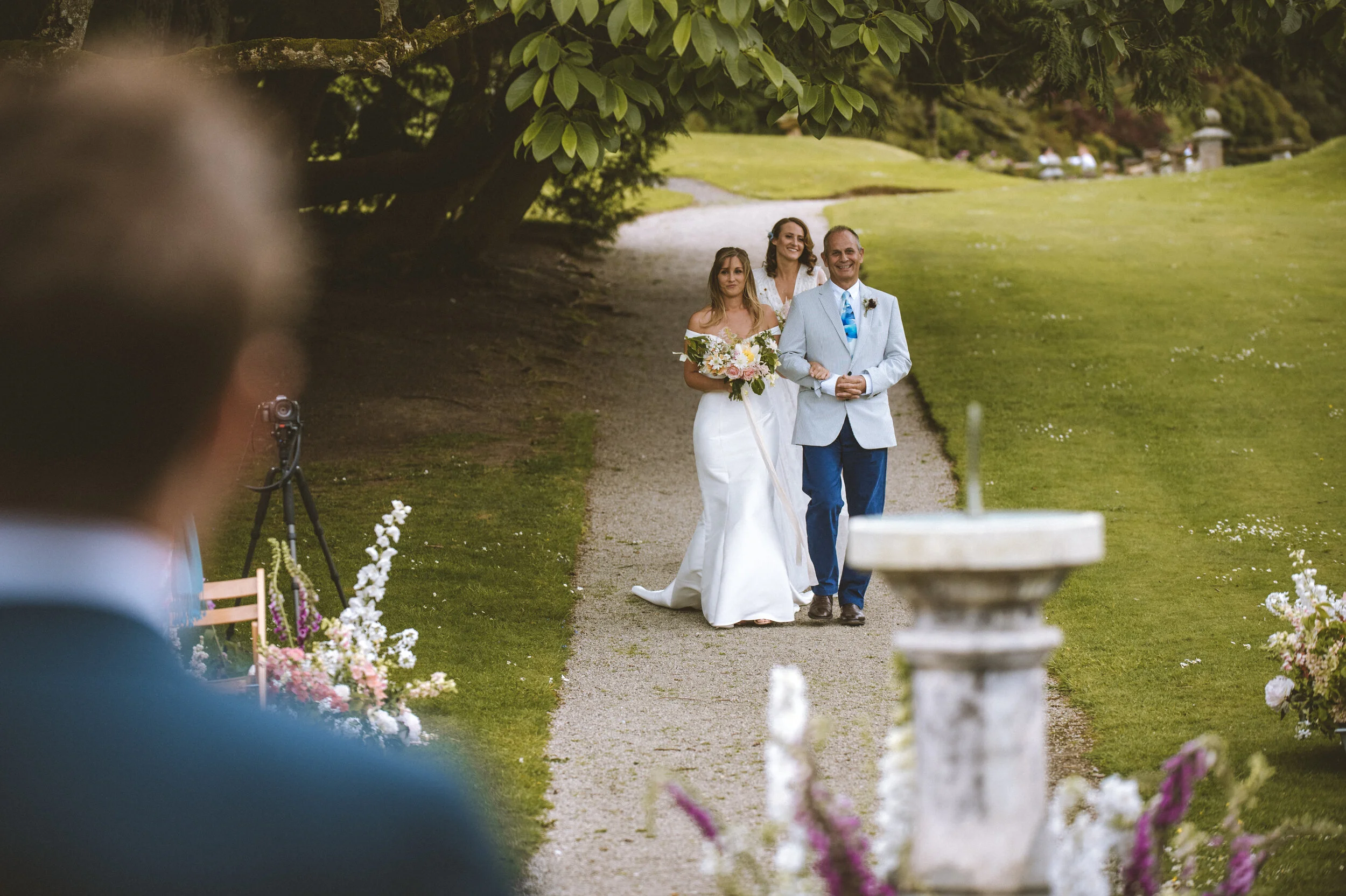 A bride walking down a gravel path with a man, likely her father, at a wedding ceremony outdoors. The bride is in a white wedding dress carrying a bouquet, and the man is in a light grey blazer with a blue tie. Two women are visible in the background