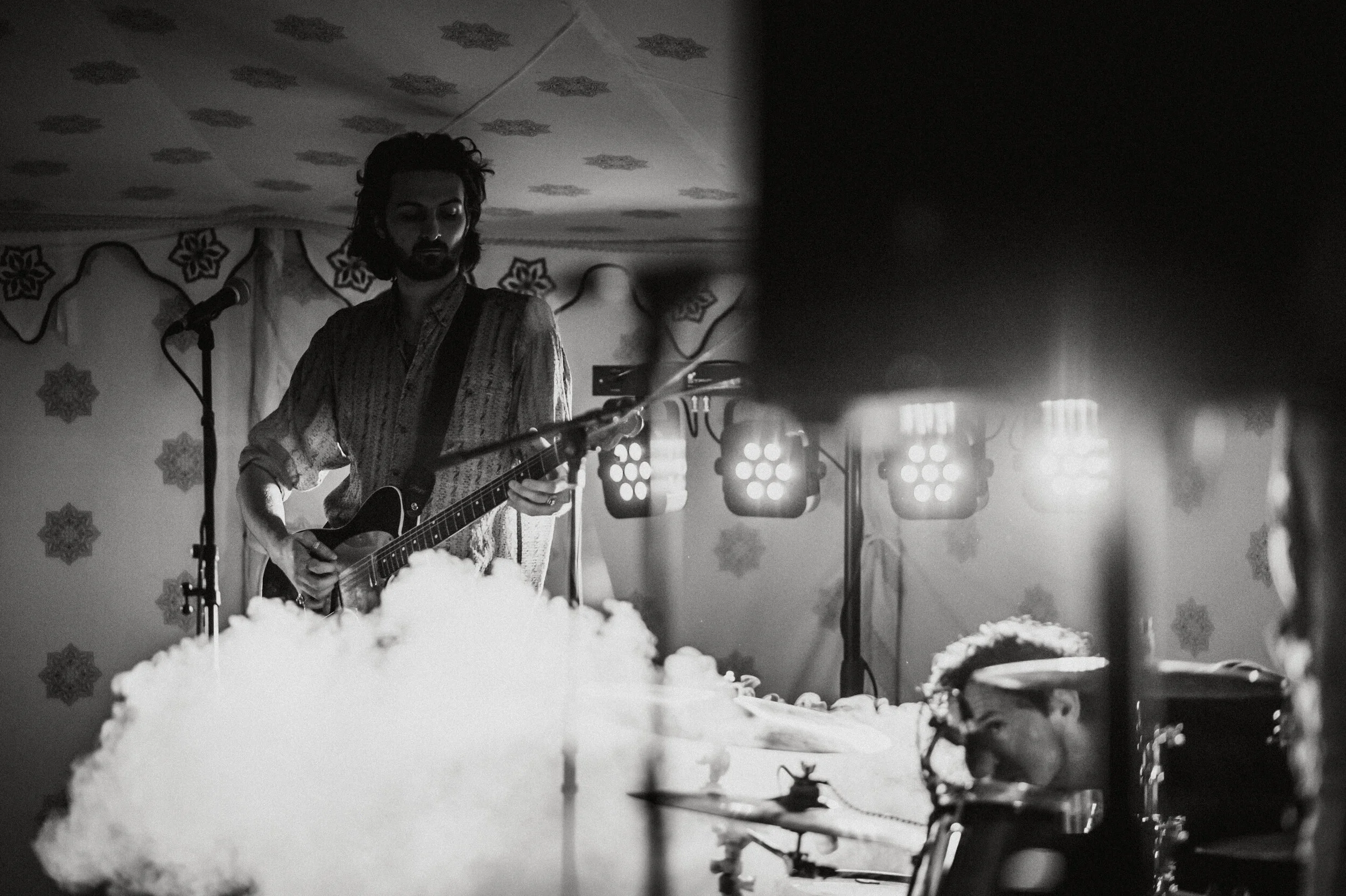 A black and white photo of a musician playing an electric guitar on stage with a microphone stand nearby, illuminated by stage lights behind him, while another band member can be seen partially on the right side.